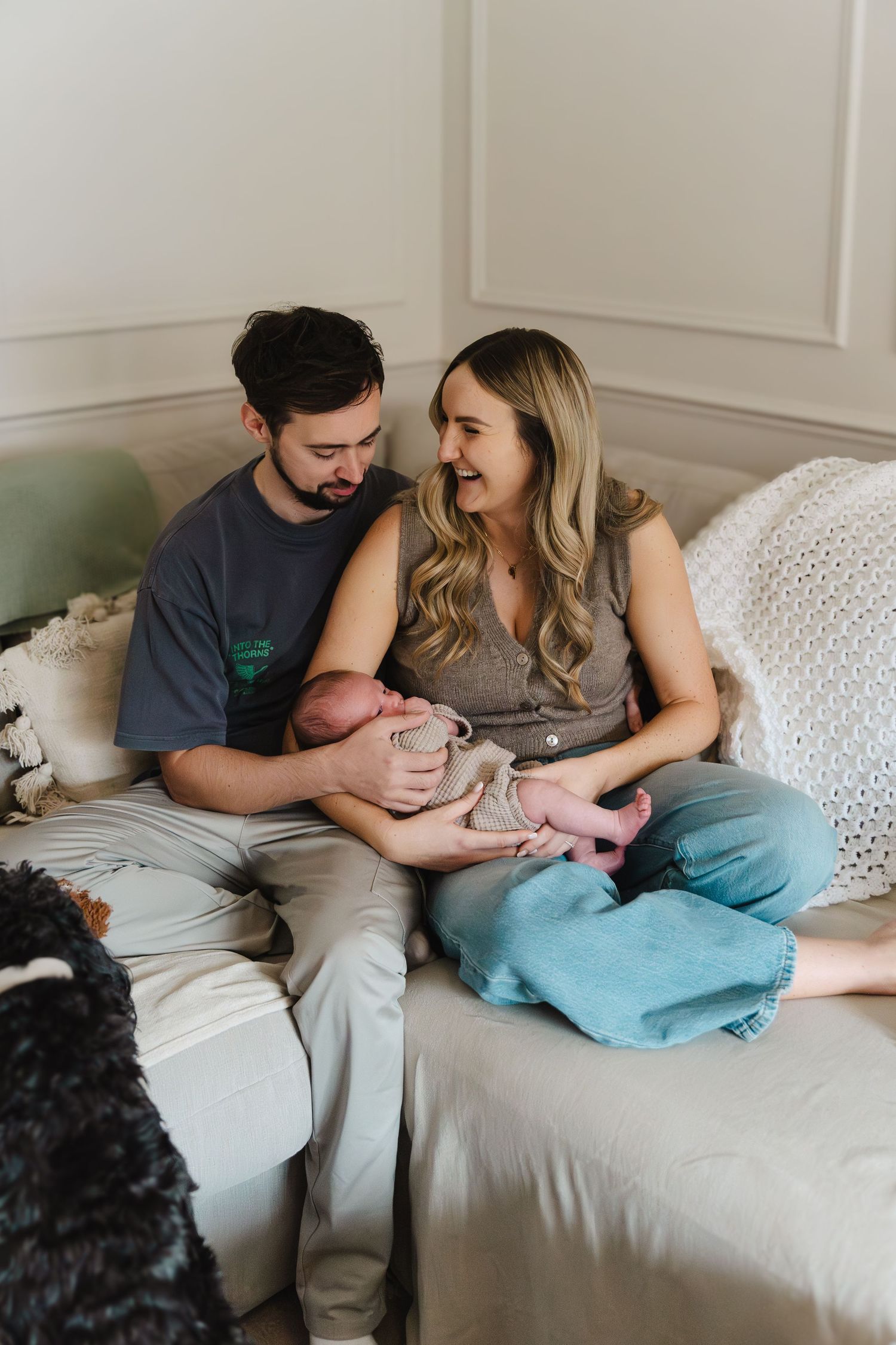 A couple sits on a white couch with their newborn baby in a series of intimate lifestyle photos.