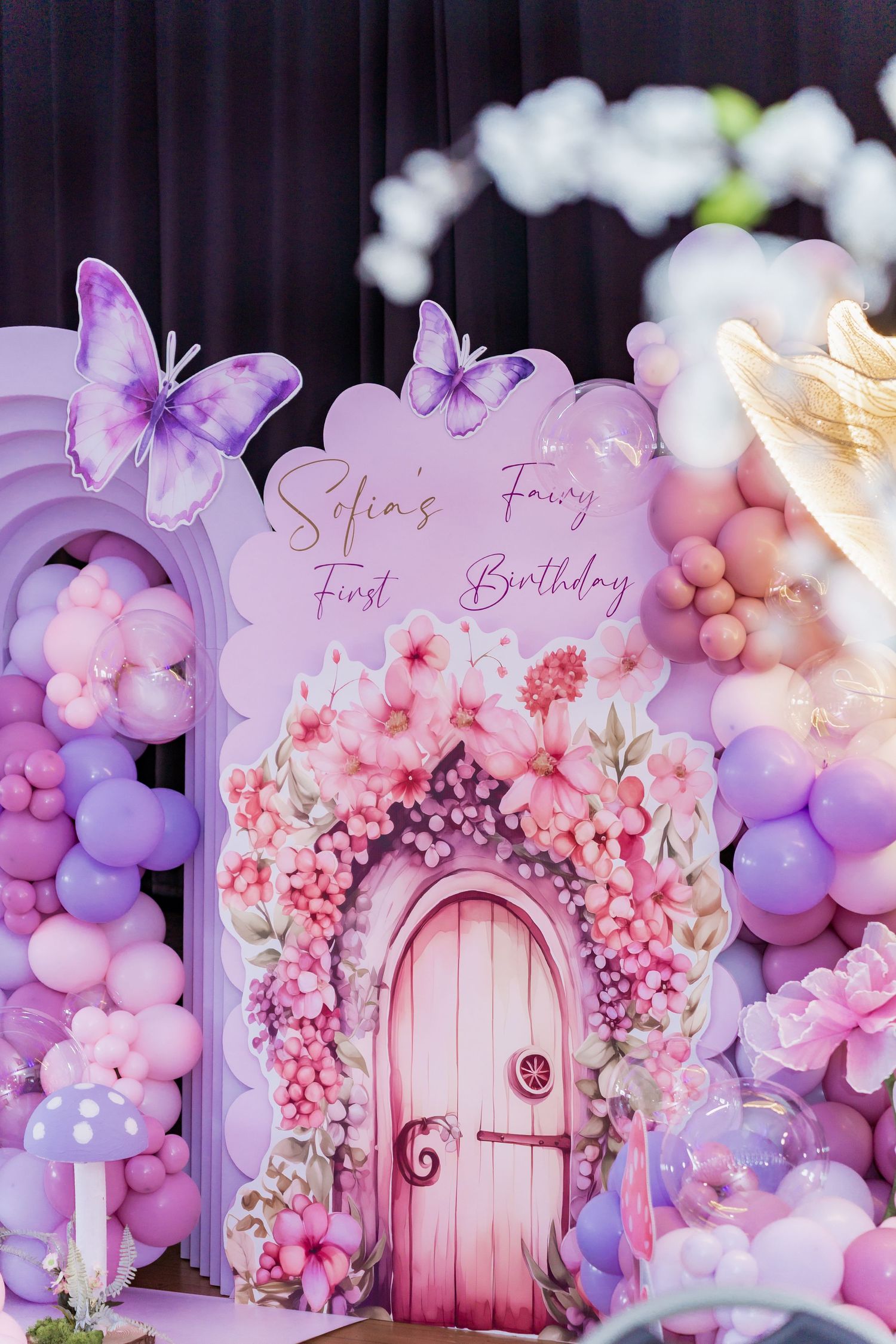 Selim's Fairy First Birthday party backdrop with pink door, floral arch, purple balloons, and butterfly decorations.