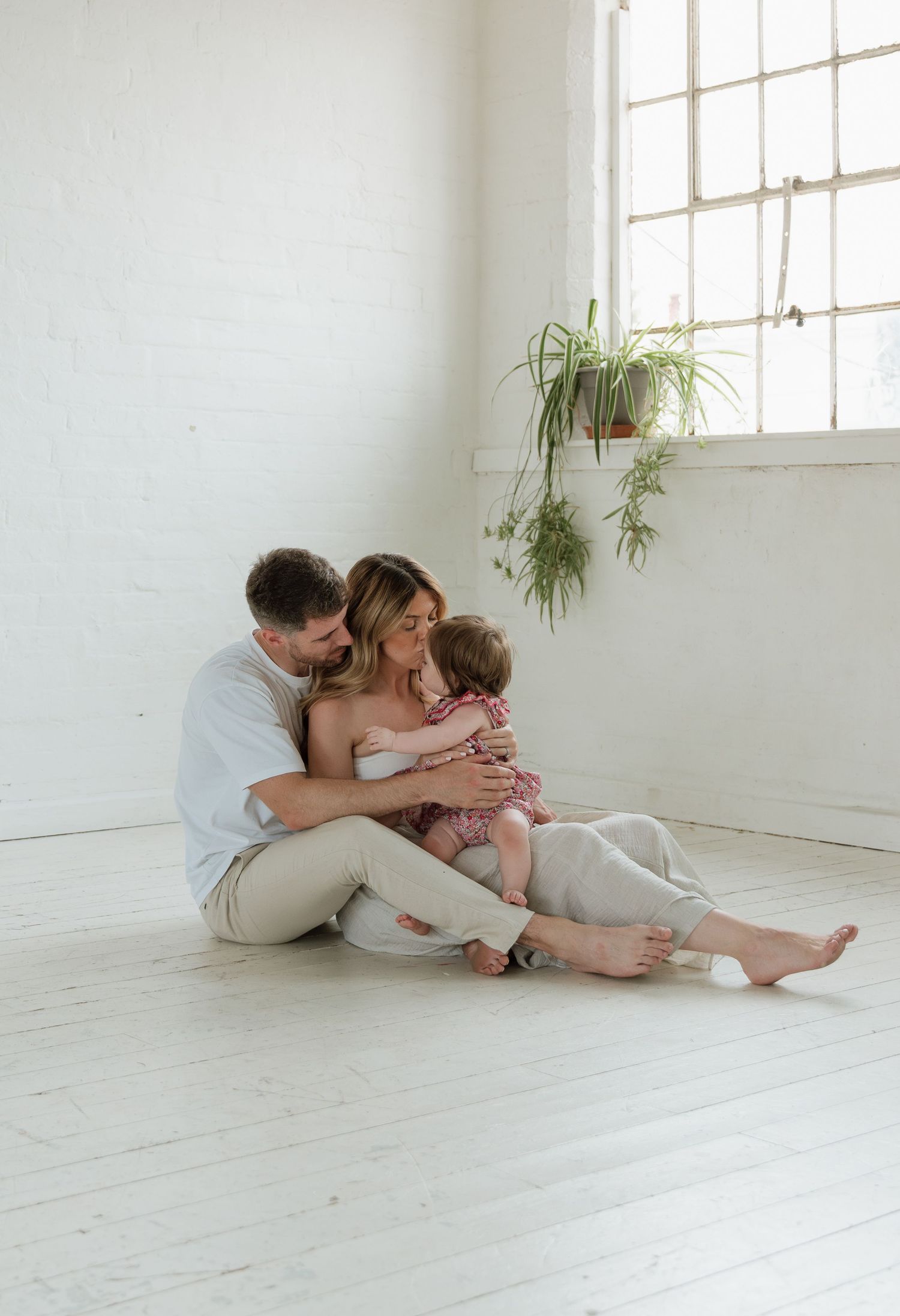 A young family sits together on a white floor, parents embracing their toddler in a bright, minimalist room with hanging plants.