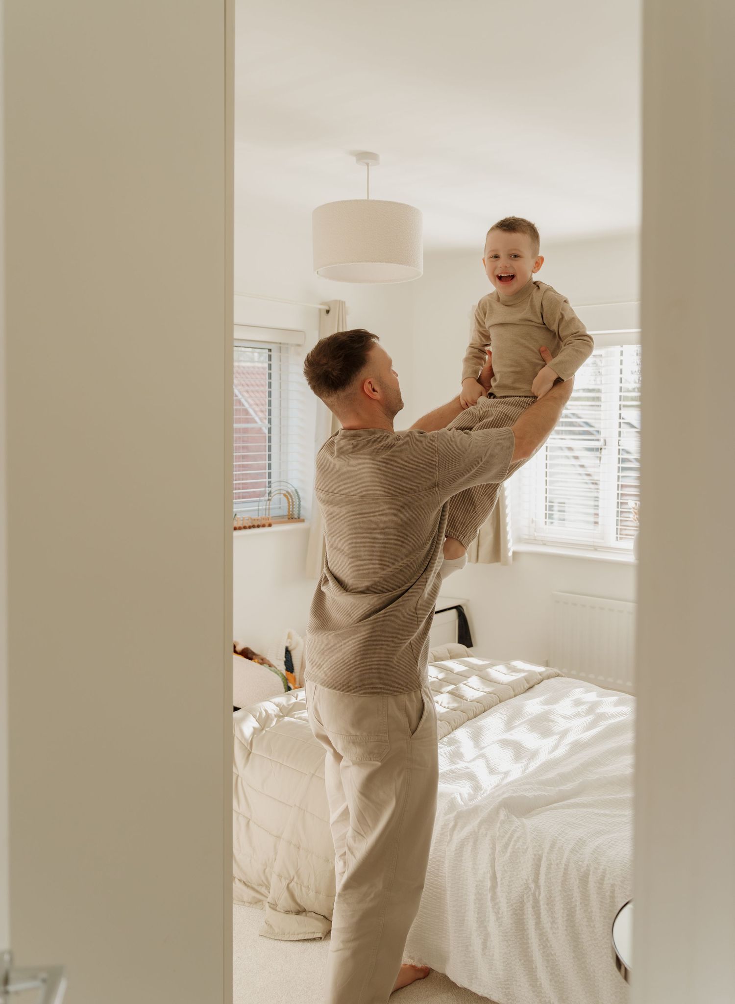 A father lifts his laughing baby in a bright, neutral-toned bedroom, both wearing matching beige outfits.