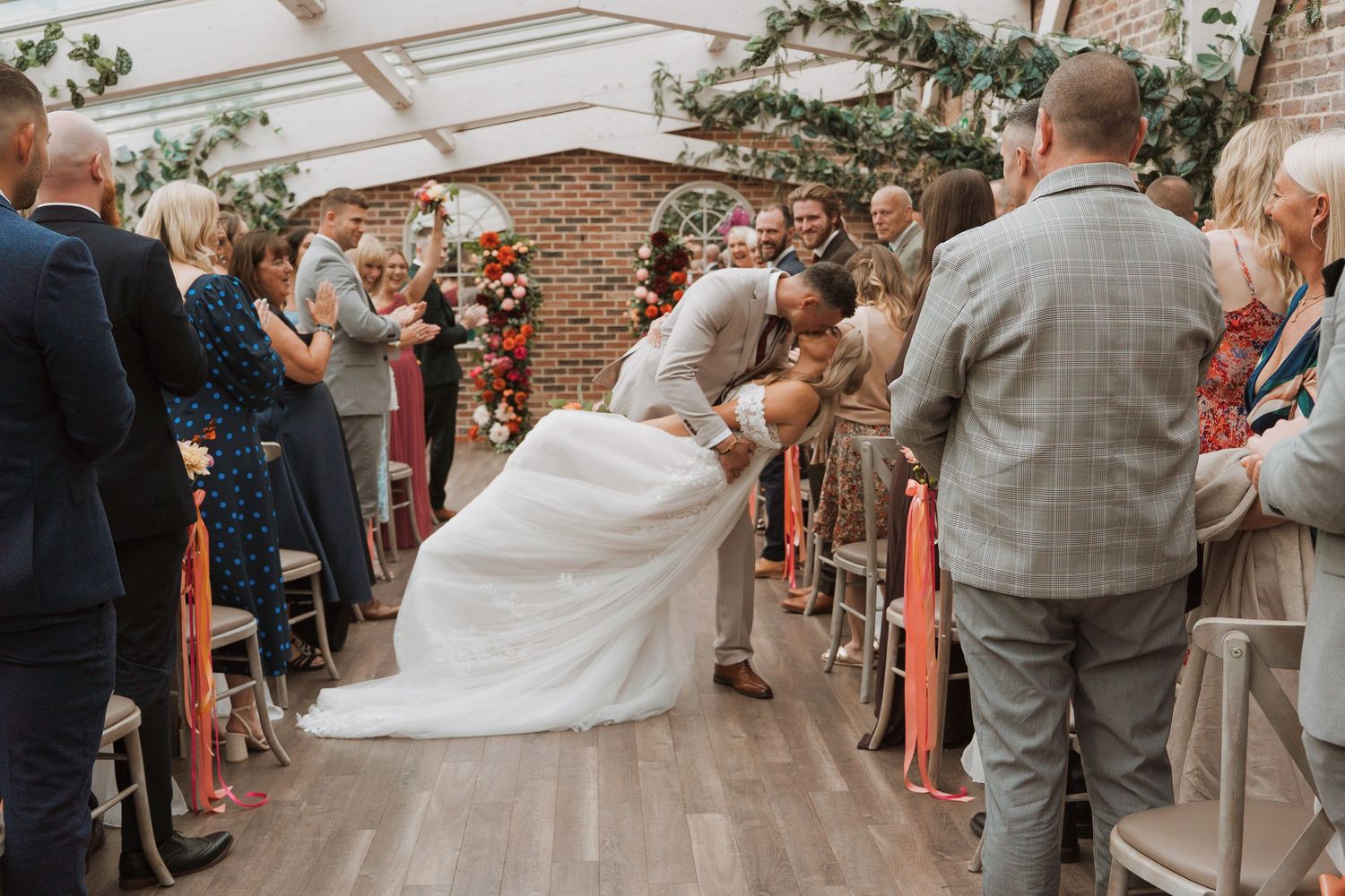 Bride and groom share a romantic dip kiss while walking down the aisle at an indoor wedding ceremony in Derbyshire, at Foxtail Barns.