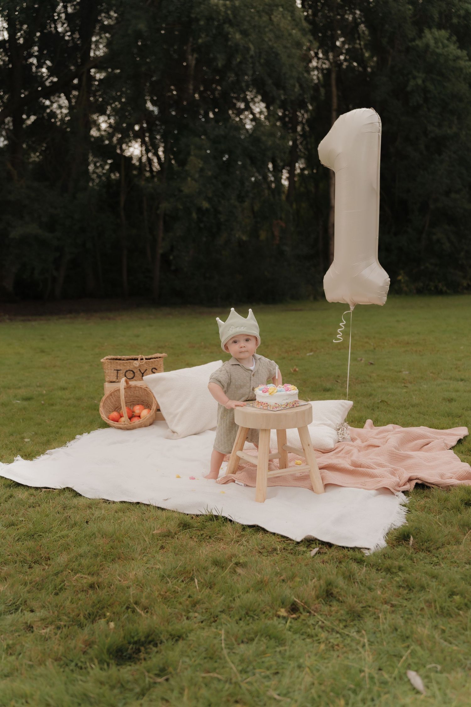 First birthday party setup outdoors in Derbyshire with white blanket, pink accents, wooden stool, cake and number one balloon.