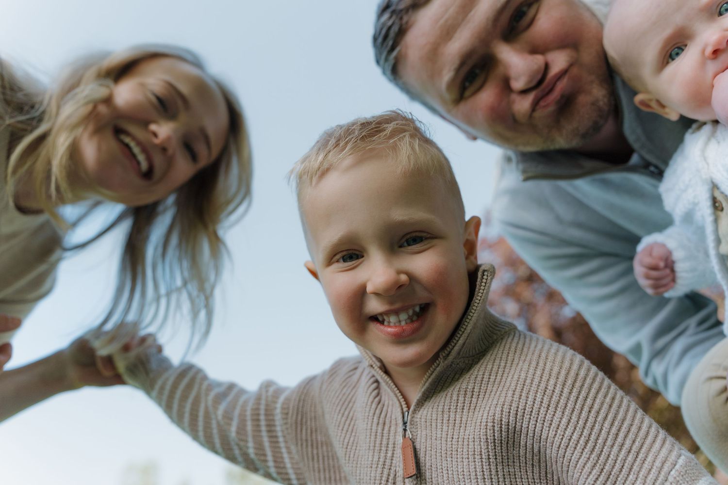 Happy family of four looking down at camera outdoors, smiling toddler boy in foreground with parents and baby.