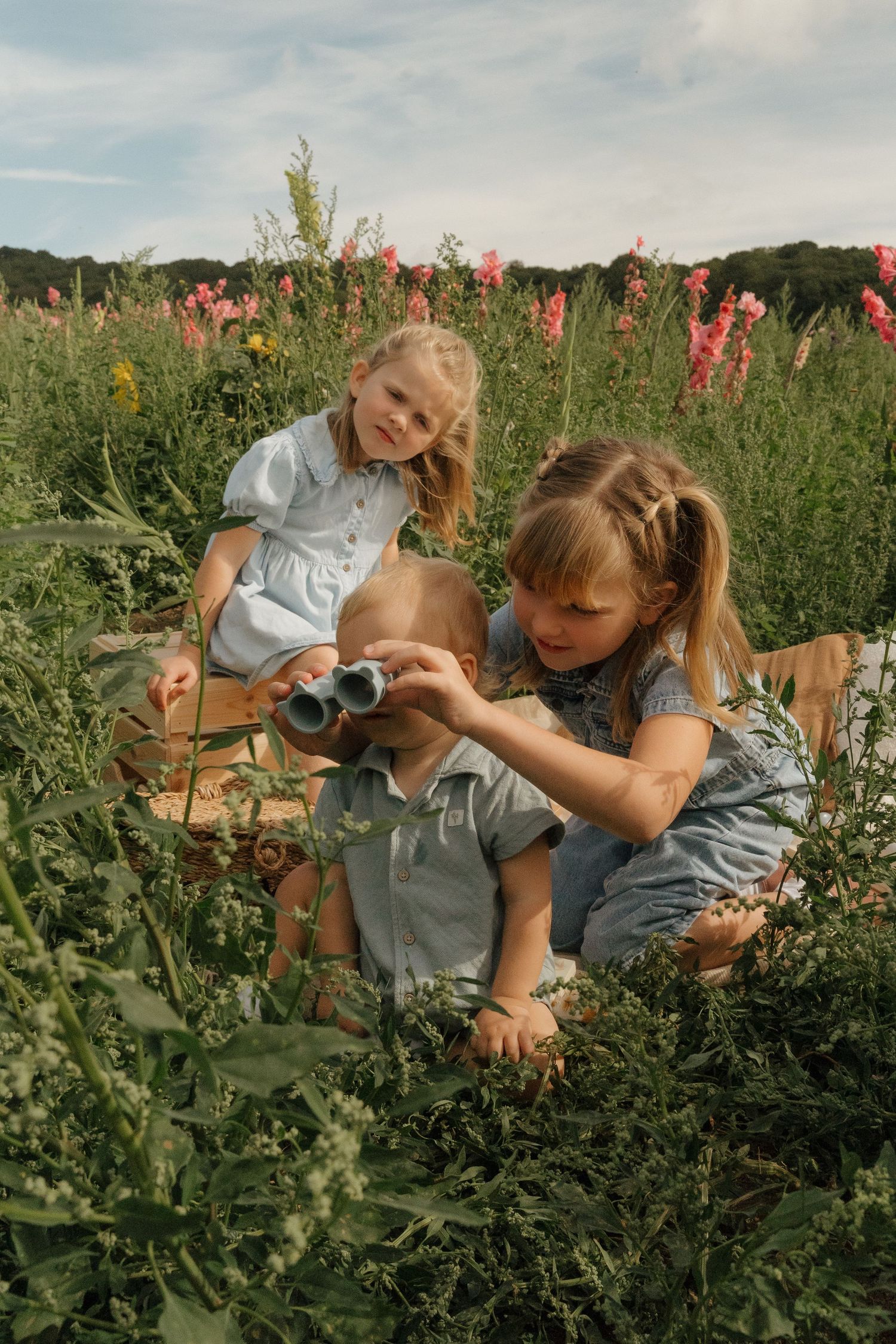 Three children playing with binoculars in a wildflower field on a sunny day.