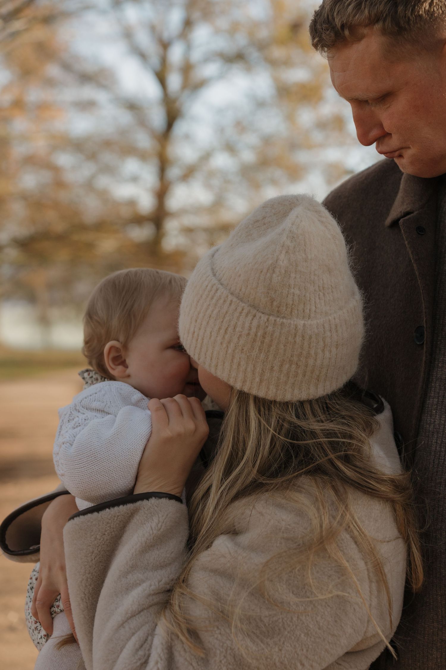 A tender family moment outdoors in autumn as a mother in a beige beanie holds her baby close while the father looks on.