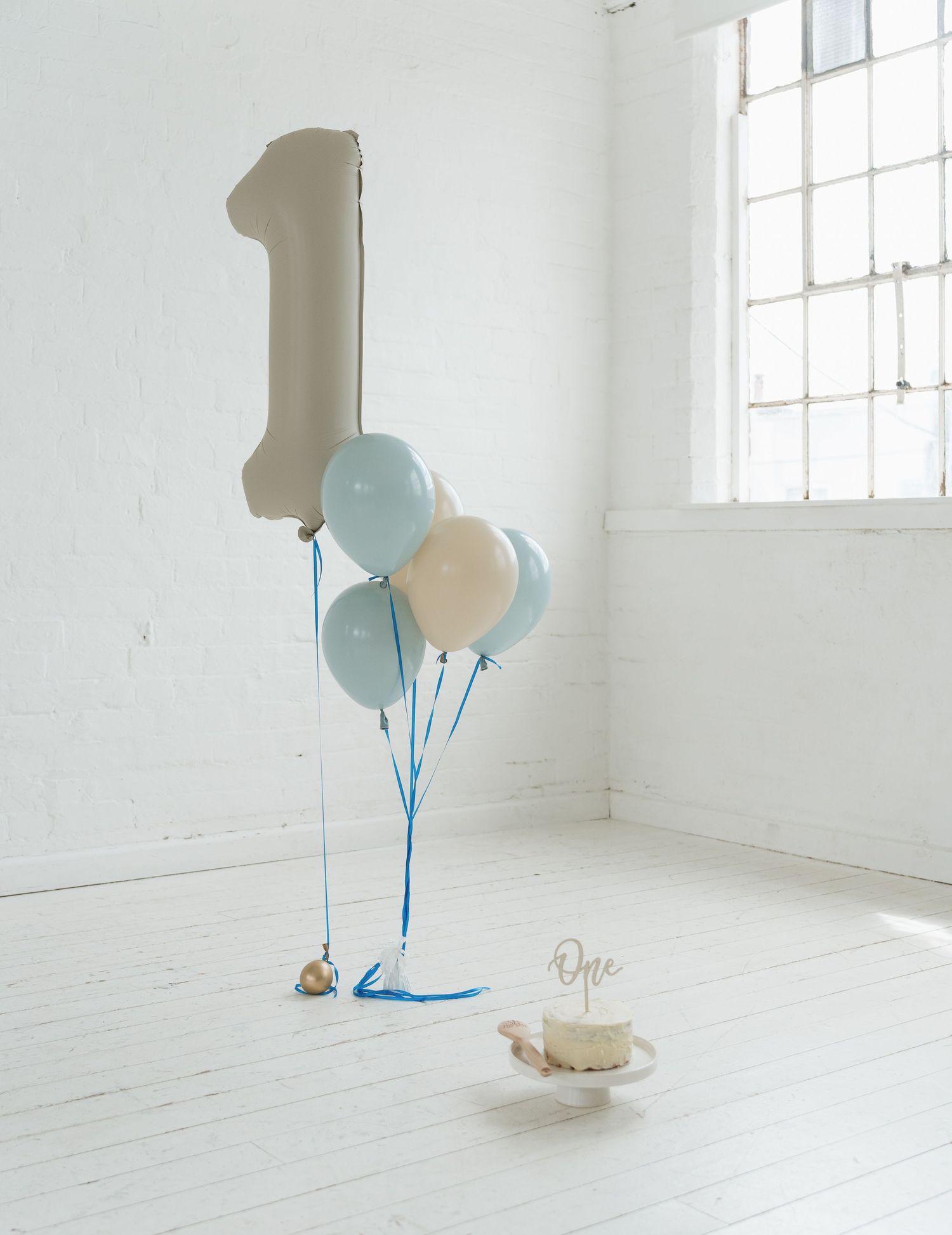 A first birthday setup with a number one balloon, blue and cream balloons, and a small smash cake on a white floor.