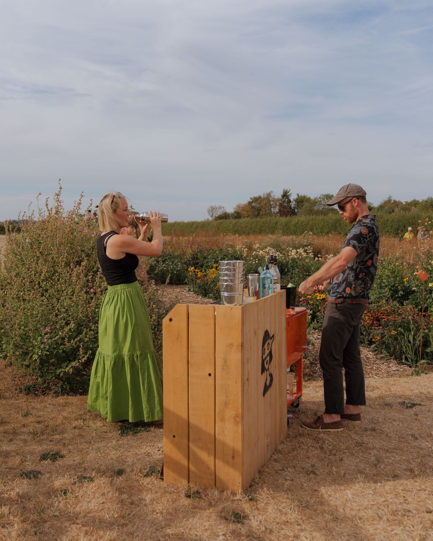 Woman in green skirt drinks from cup while man browses outdoor wooden bar stand surrounded by wildflowers.