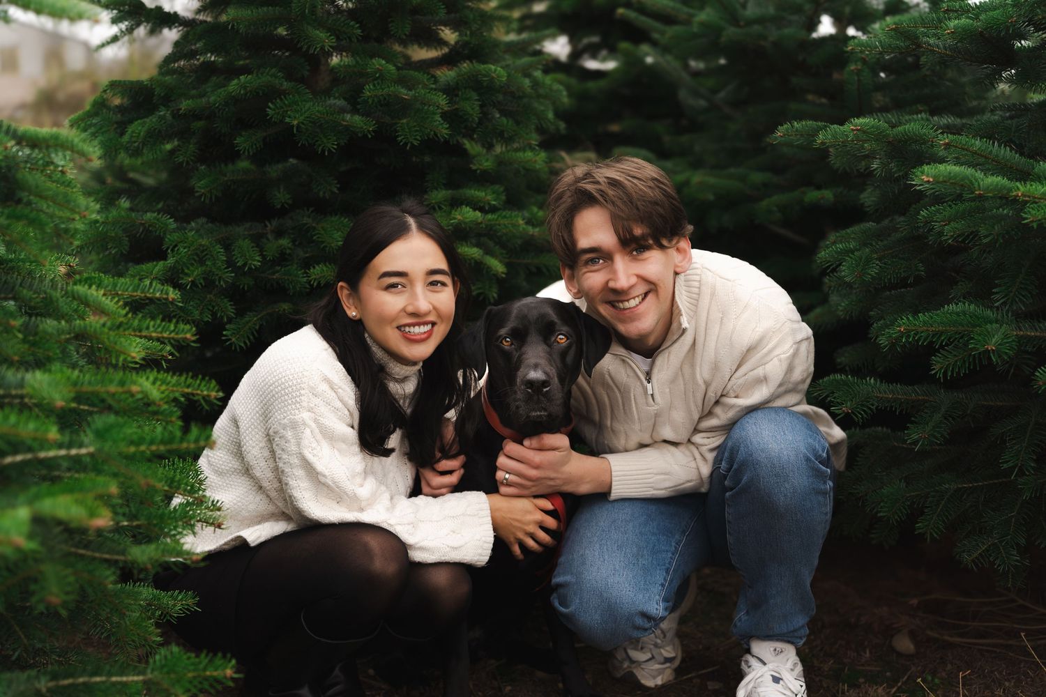 A couple sits together and laughs while taking photos in a garden with evergreen trees.