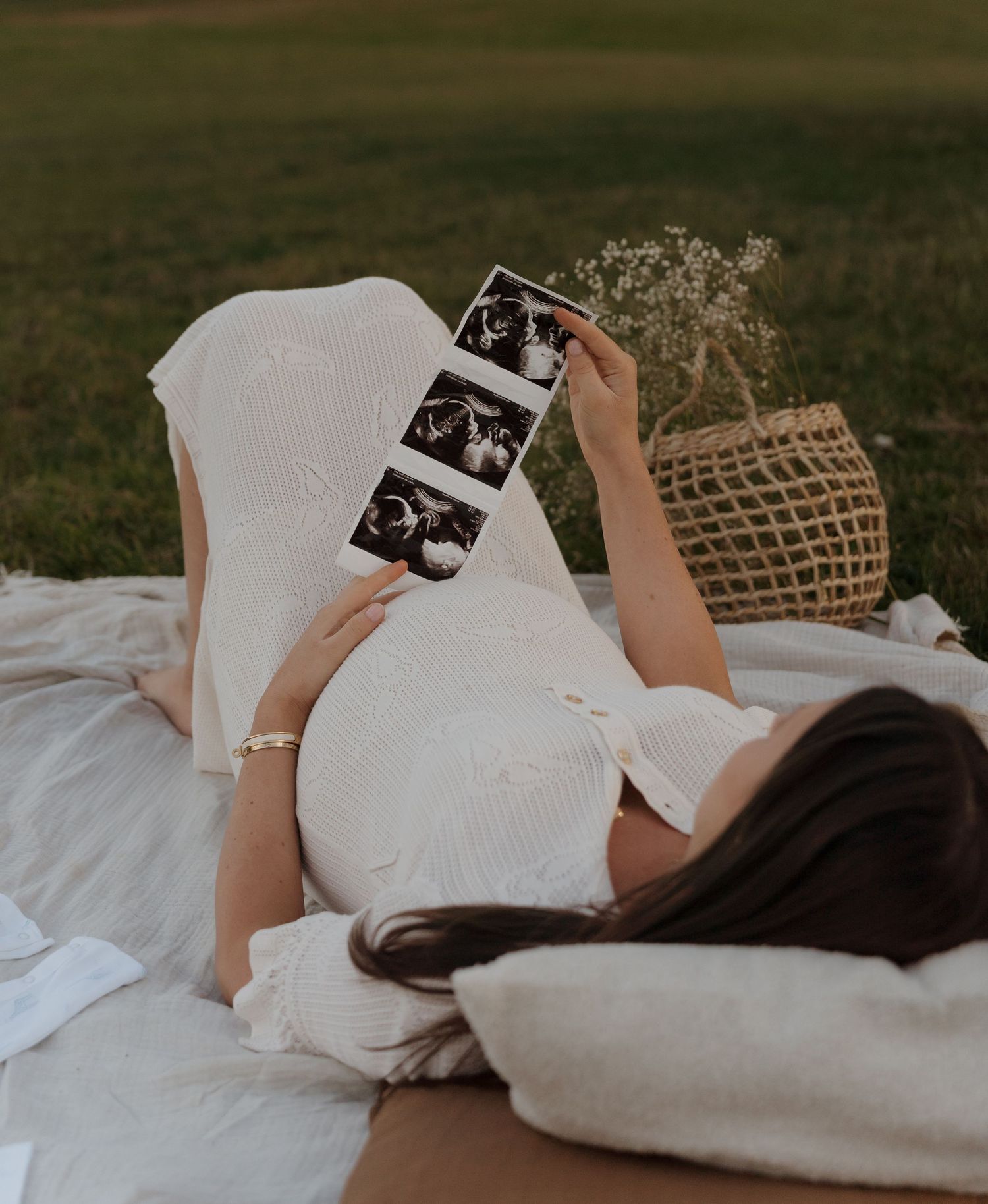 Pregnant woman in white dress lying on picnic blanket holding ultrasound photos in a grassy outdoor setting in Derbyshire.