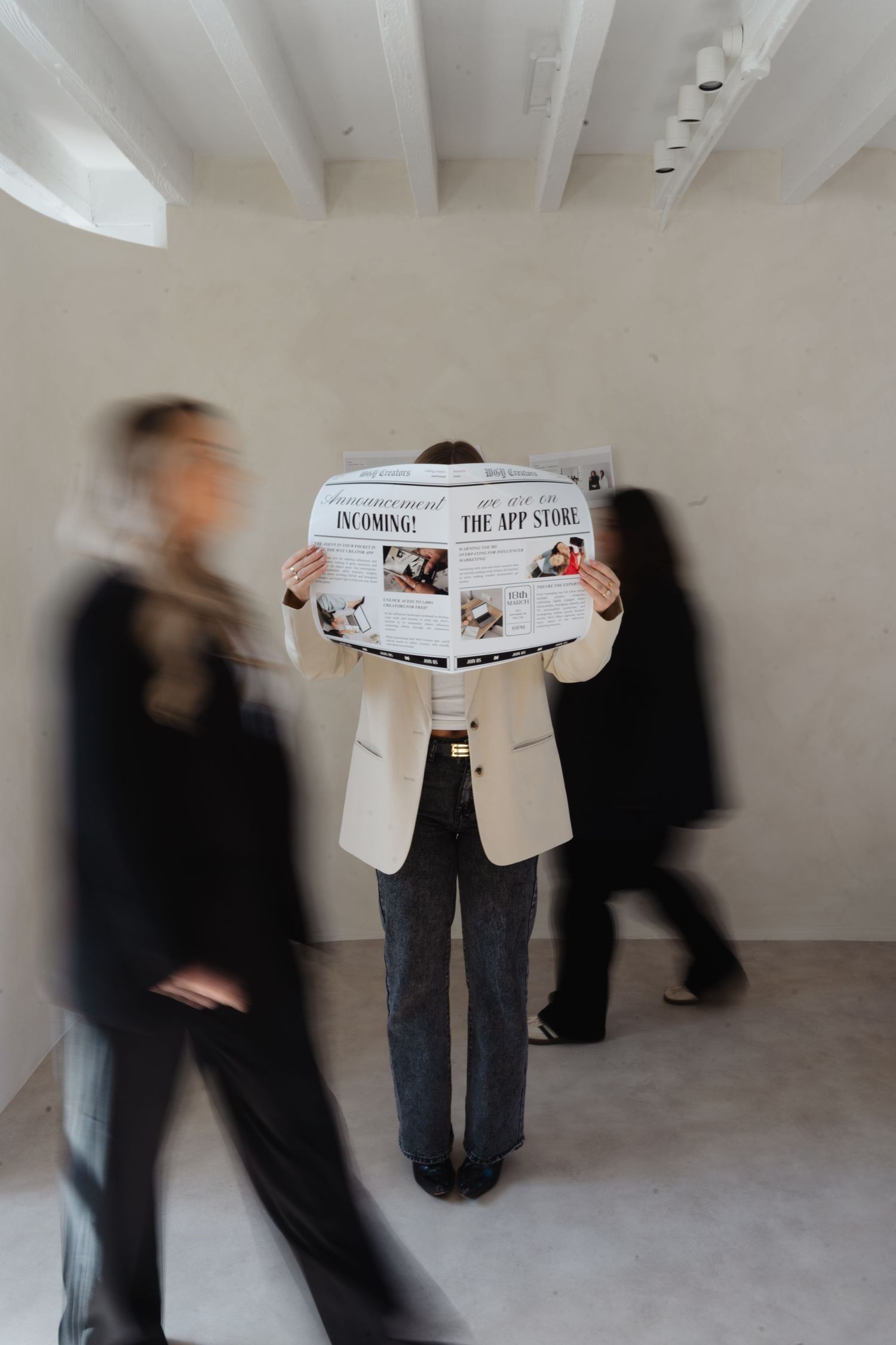 Two figures in black and white clothing perform a blurred motion sequence while holding a newspaper in an empty gallery space.