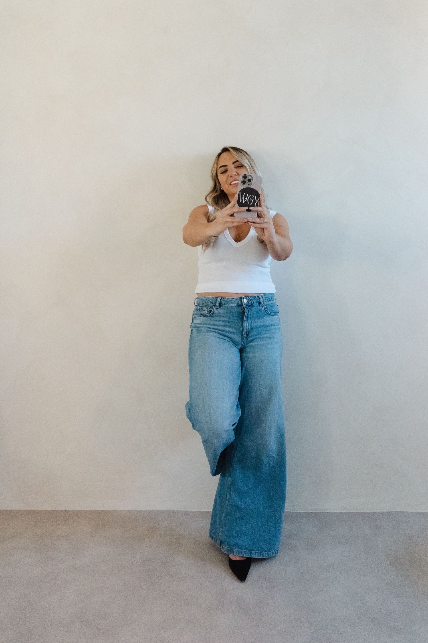 A person in flared jeans and white top taking a photo with a mobile phone against a white wall.