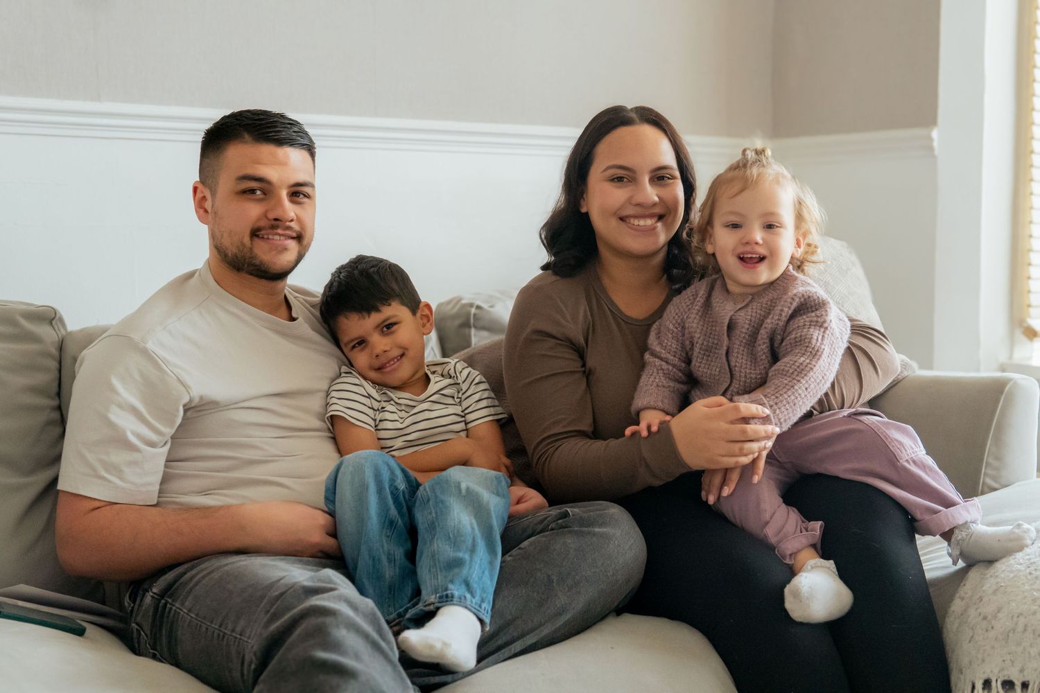 A family lounges together on a gray couch in a bright living room during a casual moment at home.