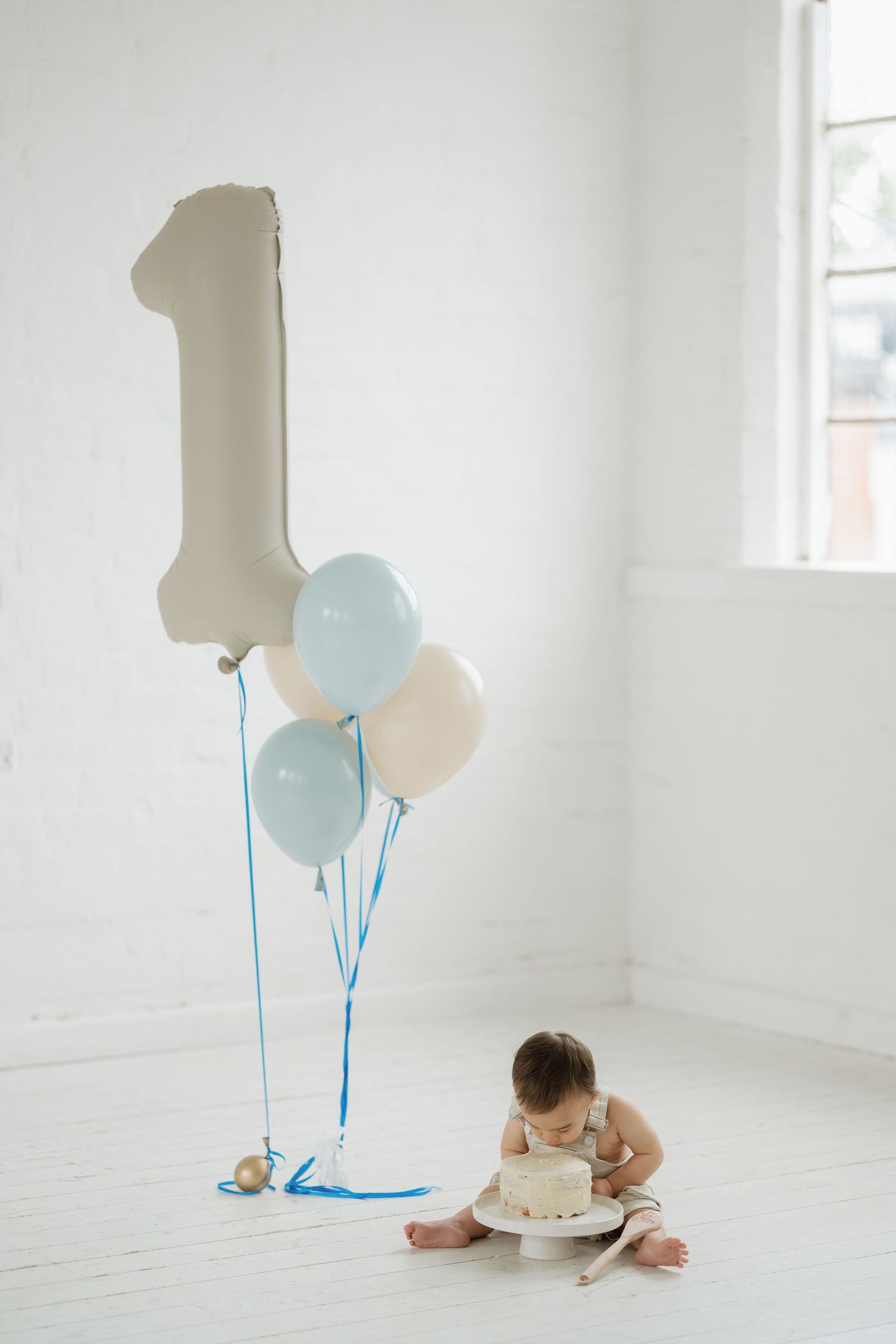 Baby enjoying first birthday cake smash with blue and white balloons and number one balloon in white room.