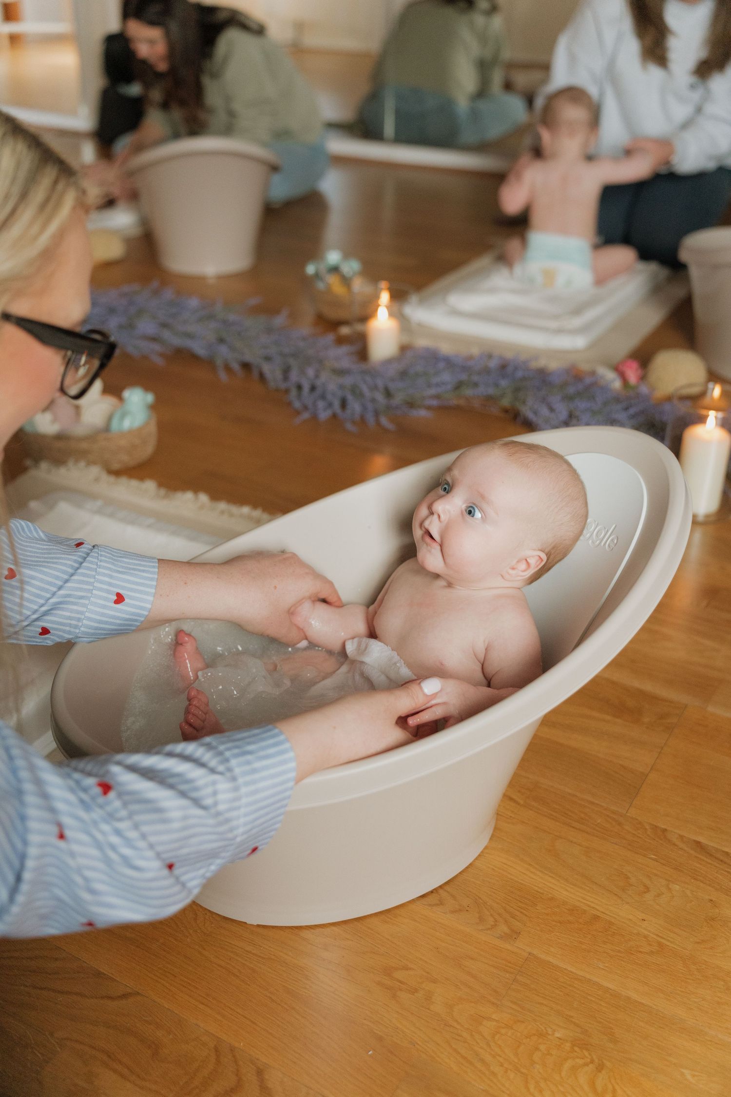 A smiling baby enjoys a bath in a small tub while a woman in a striped shirt gently holds them during a baby class.