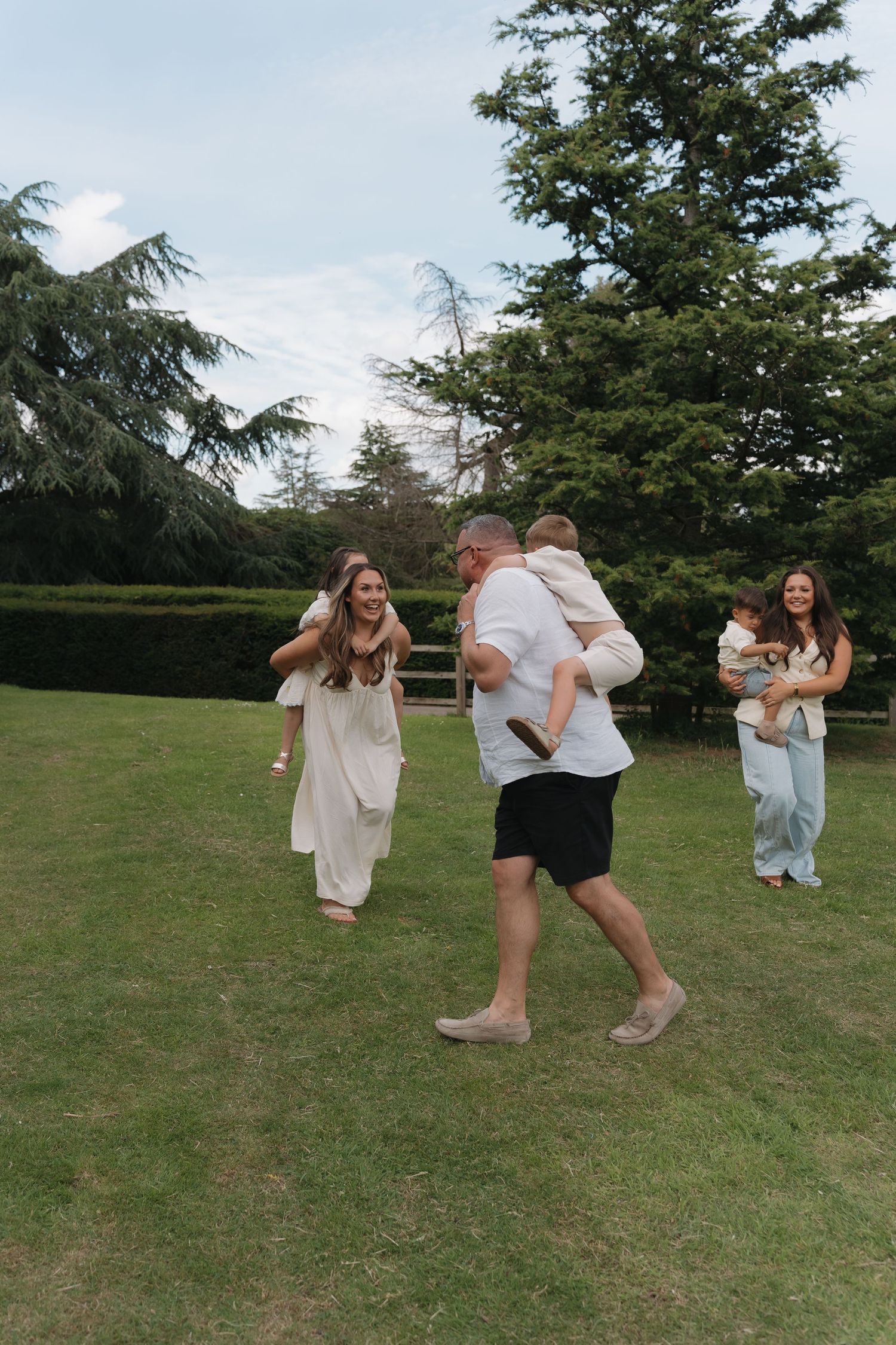 Two families playing outdoors on green lawn with large evergreen trees, parents carrying young children in casual summer attire.