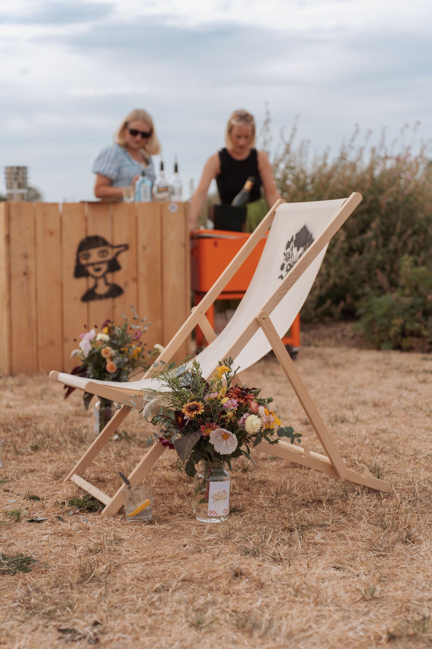 Wooden deck chair with floral bouquet at outdoor festival bar with two women in background.