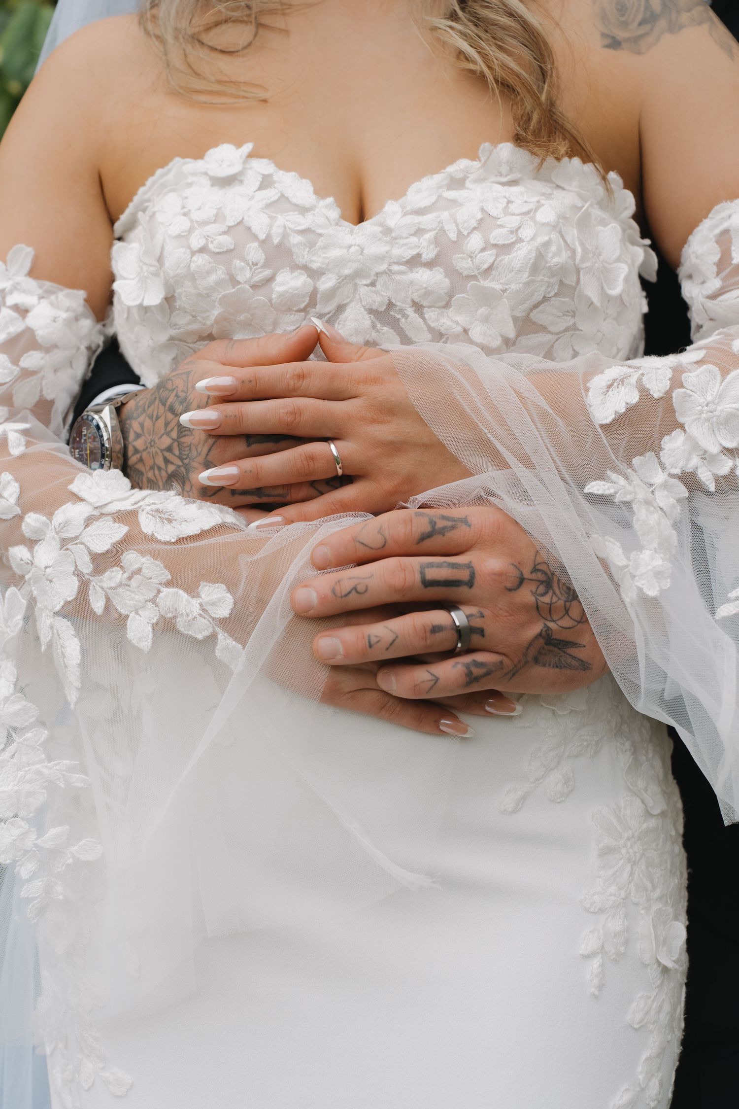 Close-up of tattooed hands resting on a white wedding dress with delicate floral appliques and lace details.