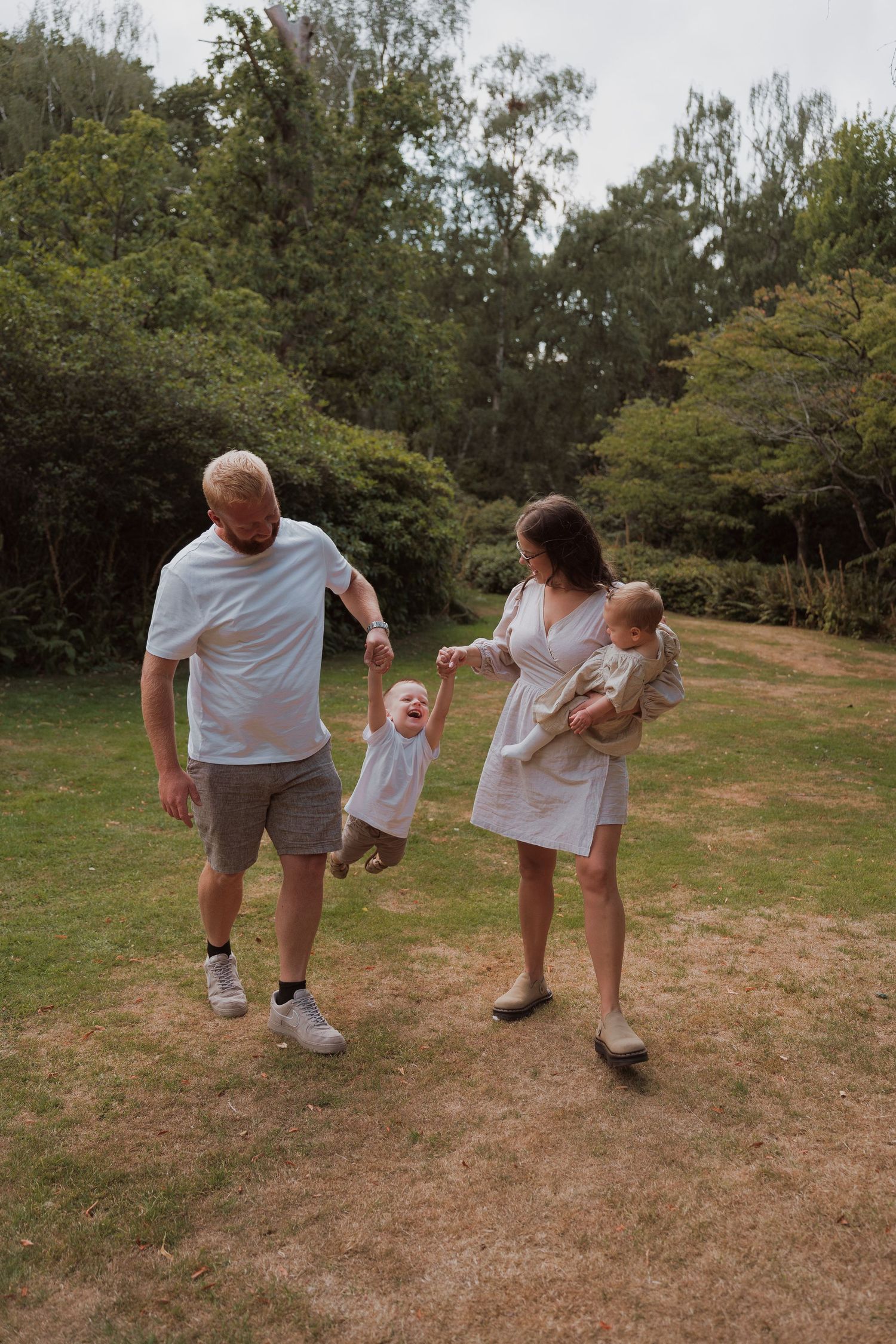 A family enjoying a sunny day outdoors, with parents swinging a toddler while holding a baby in a lush green park.