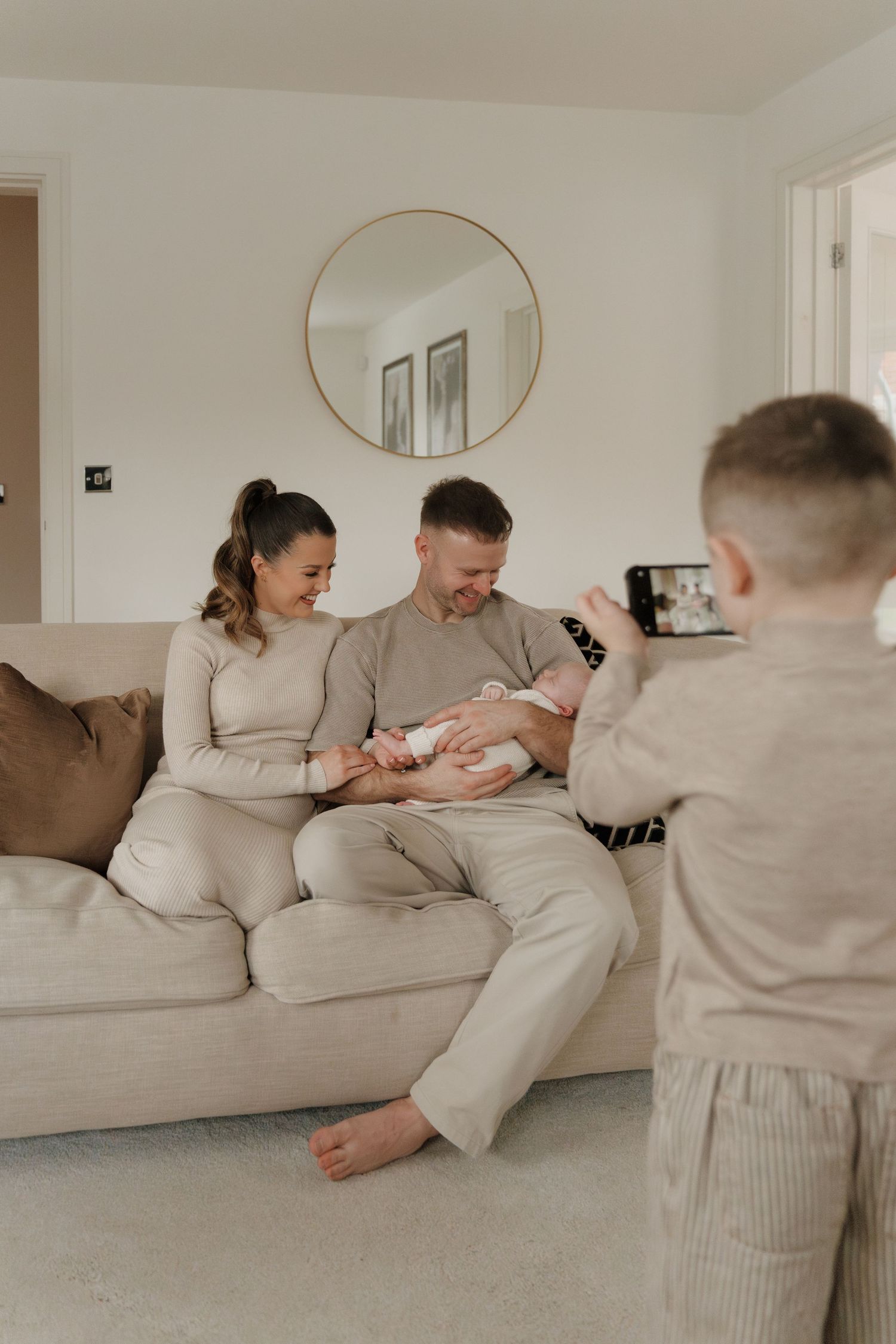 A young boy photographs his parents on a beige sofa holding their newborn baby in a bright living room.