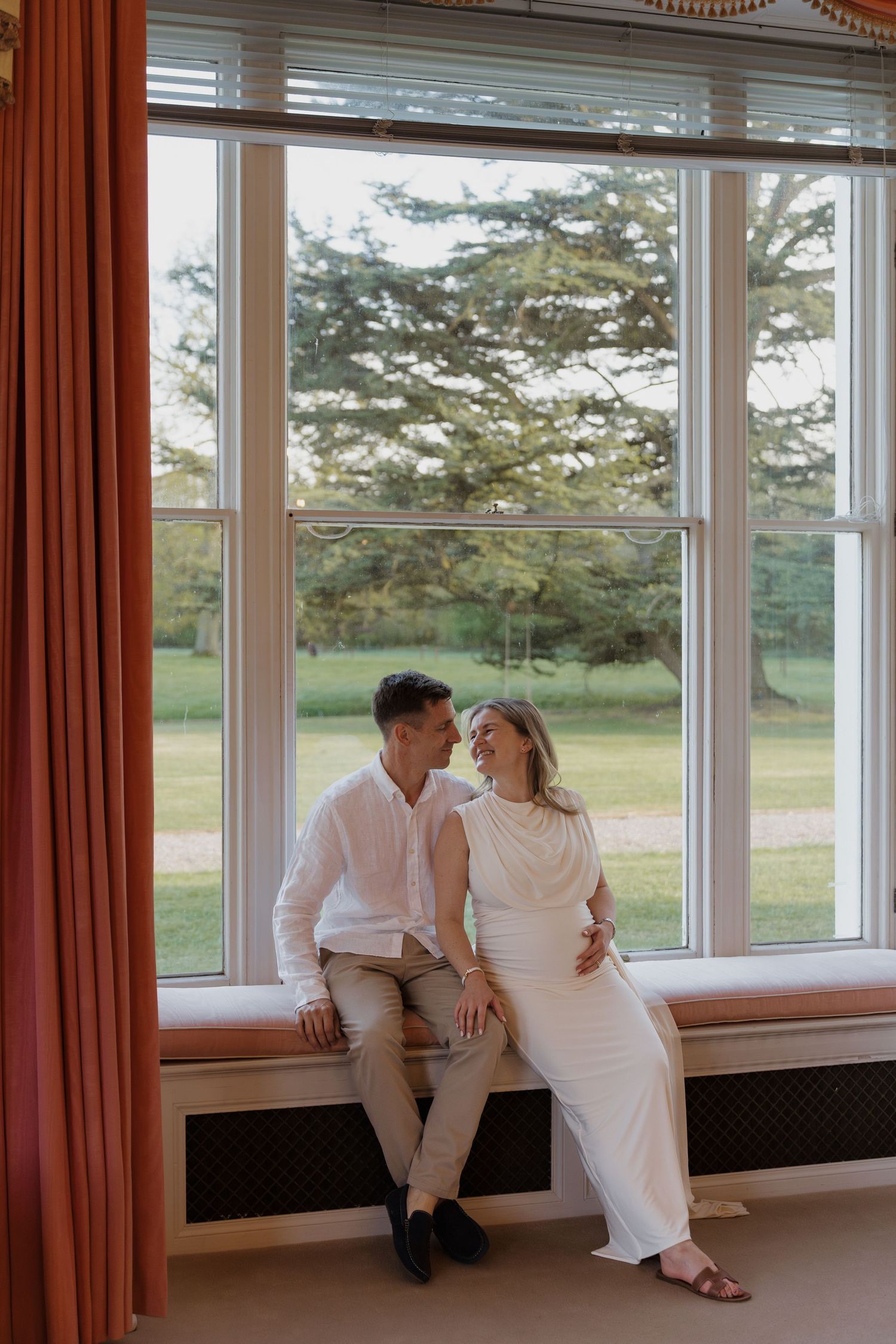 A couple sits together on a window seat in an elegant room, sharing a tender moment with a lush garden visible behind them.