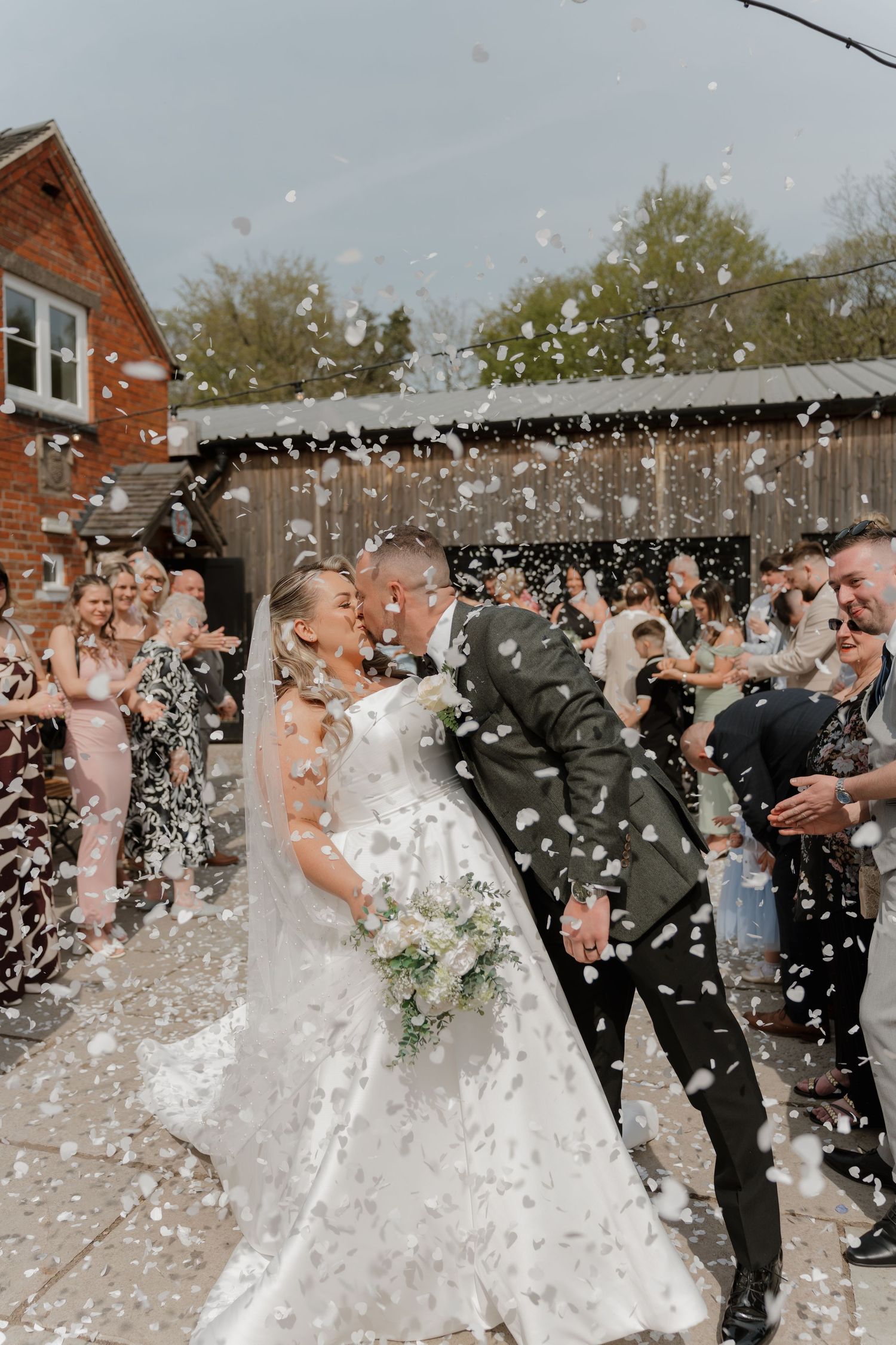Newlyweds share a kiss surrounded by white confetti, with wedding guests cheering outside a rustic brick venue.