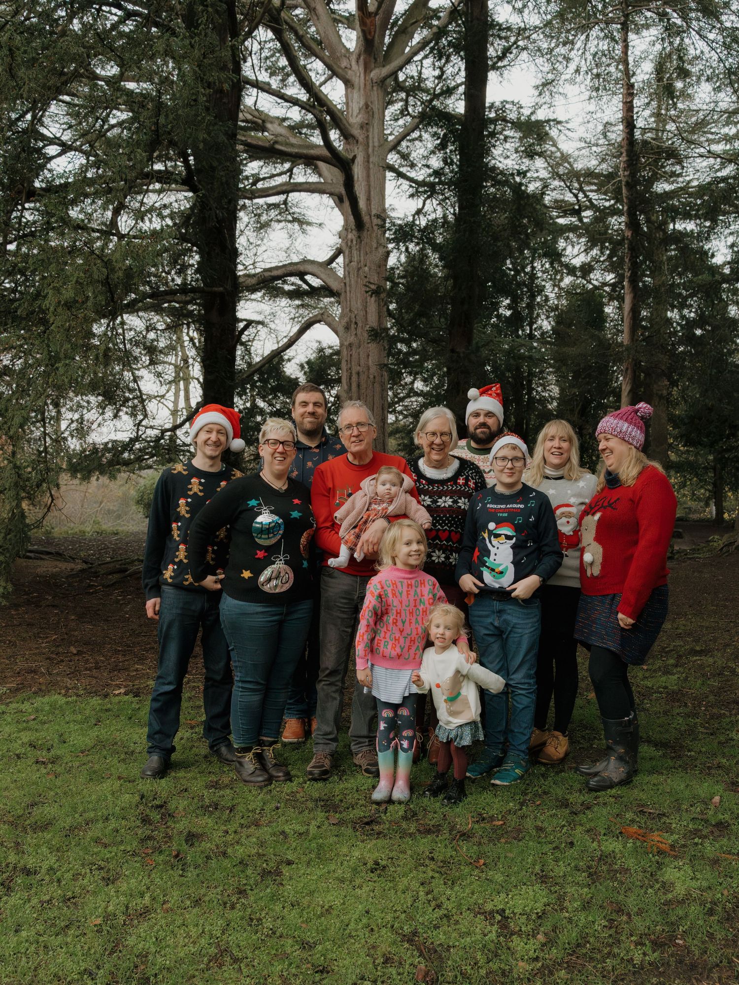Family group photo in Christmas sweaters outdoors near tall trees on a cloudy winter day.