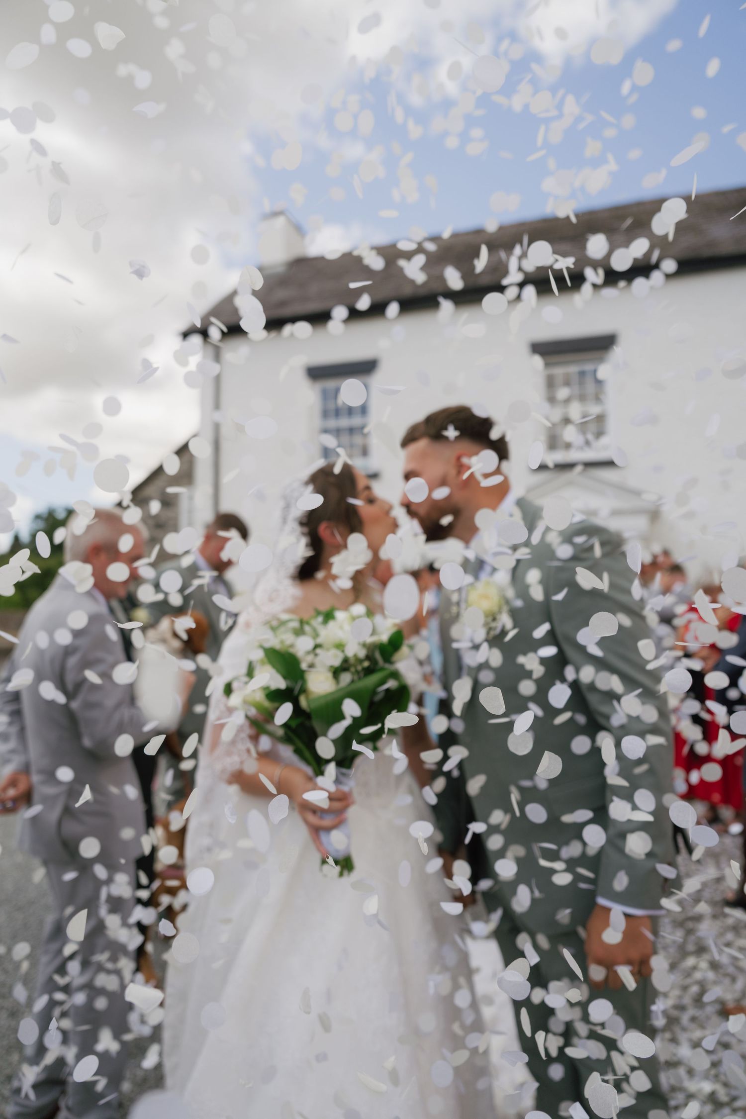 Bride and groom kissing under white confetti shower outside a white building with wedding guests.