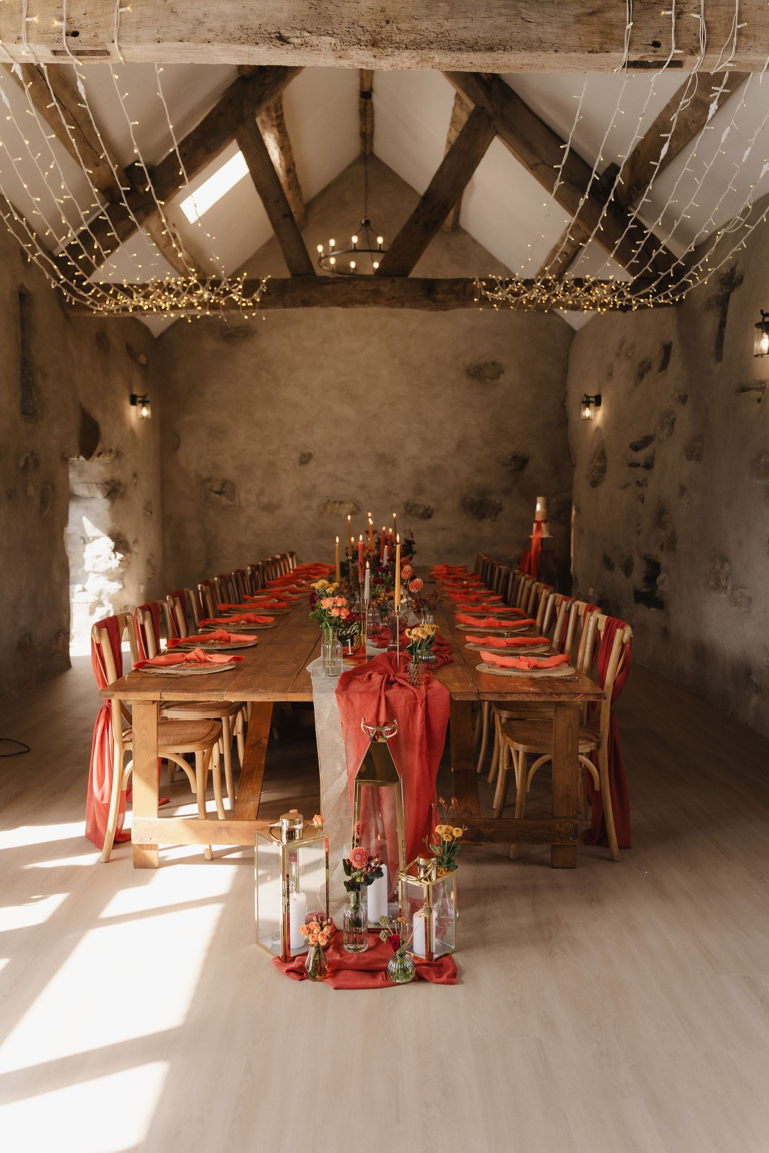 Rustic wedding reception table with red runners and string lights in a stone-walled barn venue with vaulted ceiling.
