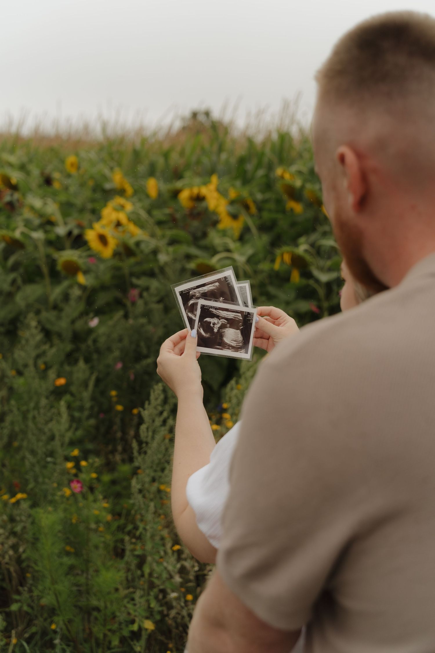 Someone in a beige suit holds up a sonogram photo in a field of sunflowers and greenery.