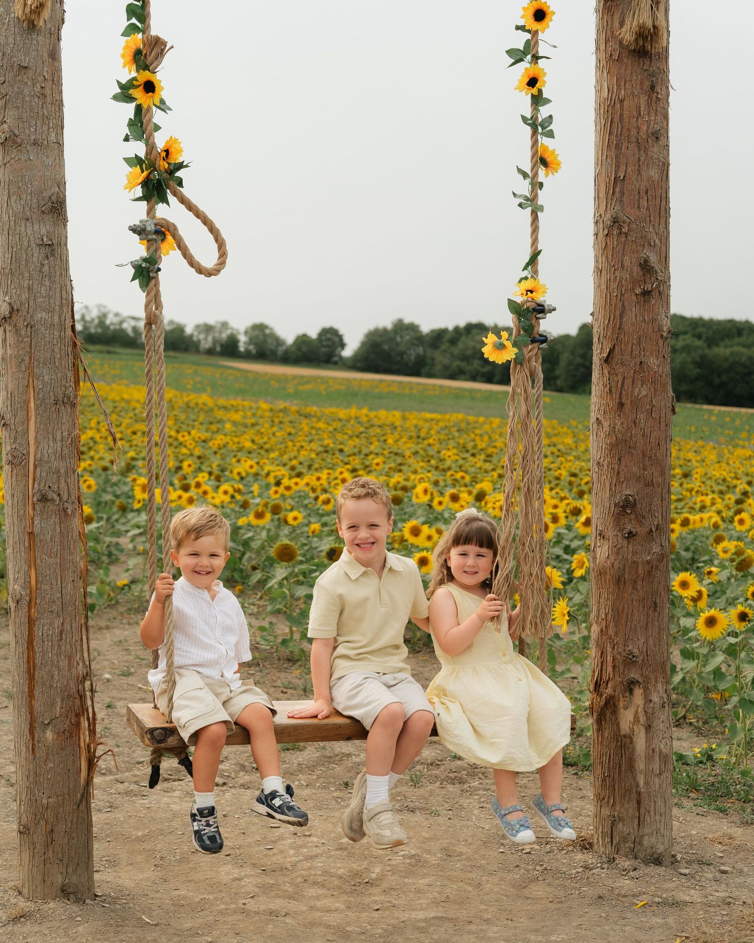 Three children sit on a rope swing decorated with sunflowers, in front of a vibrant sunflower field.