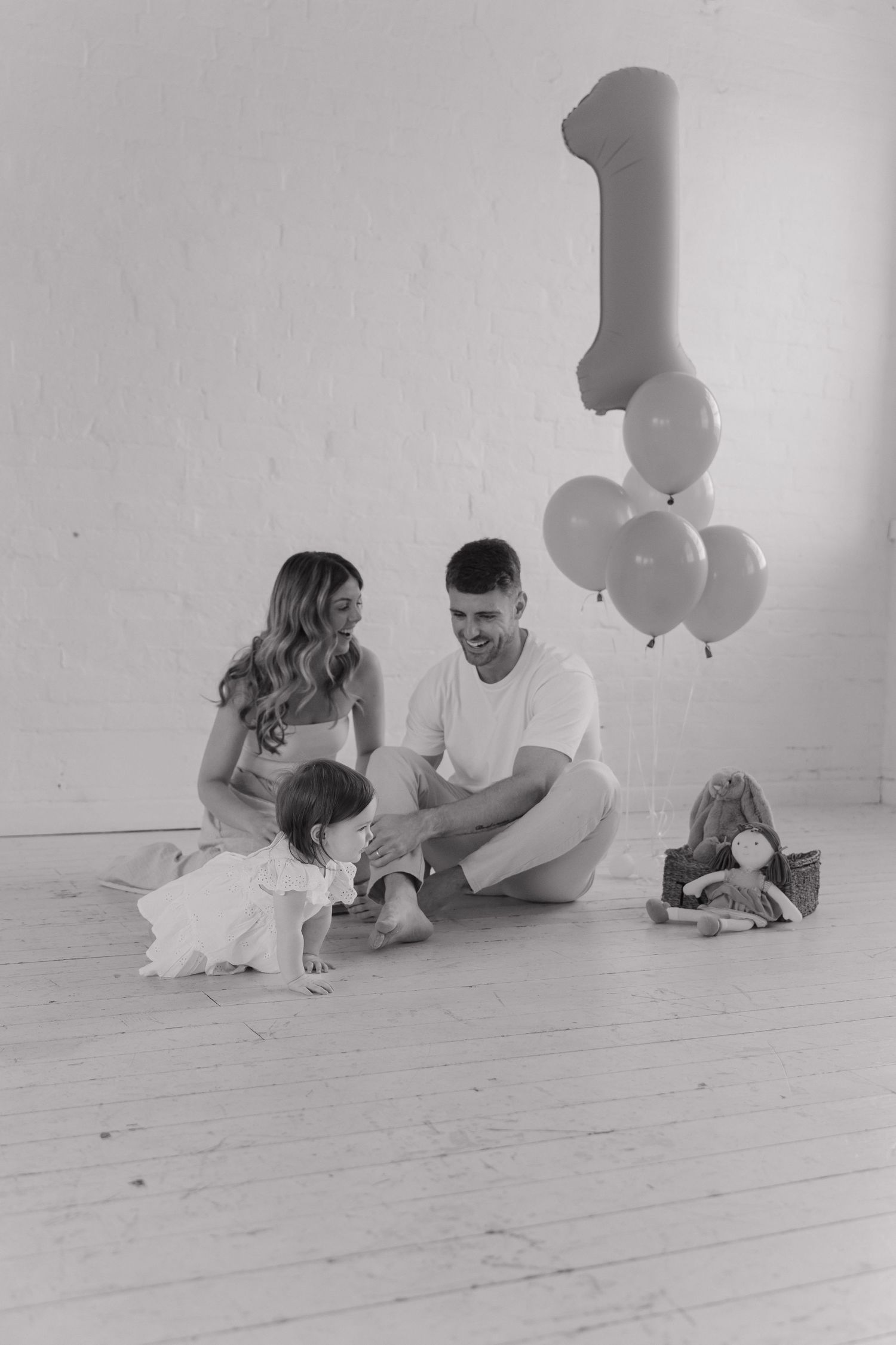 Black and white photo of a family celebrating their baby's first birthday with balloons on a white studio floor.