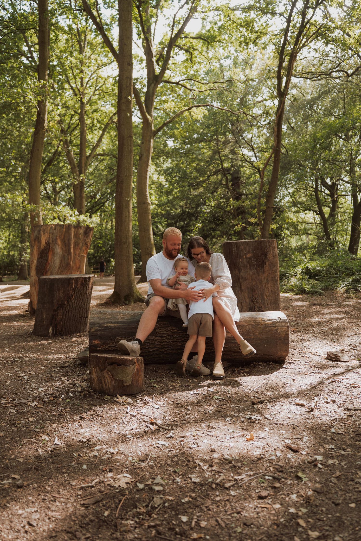 Family of three sitting on a rustic log bench in a sunlit forest, parents lovingly holding their young child.