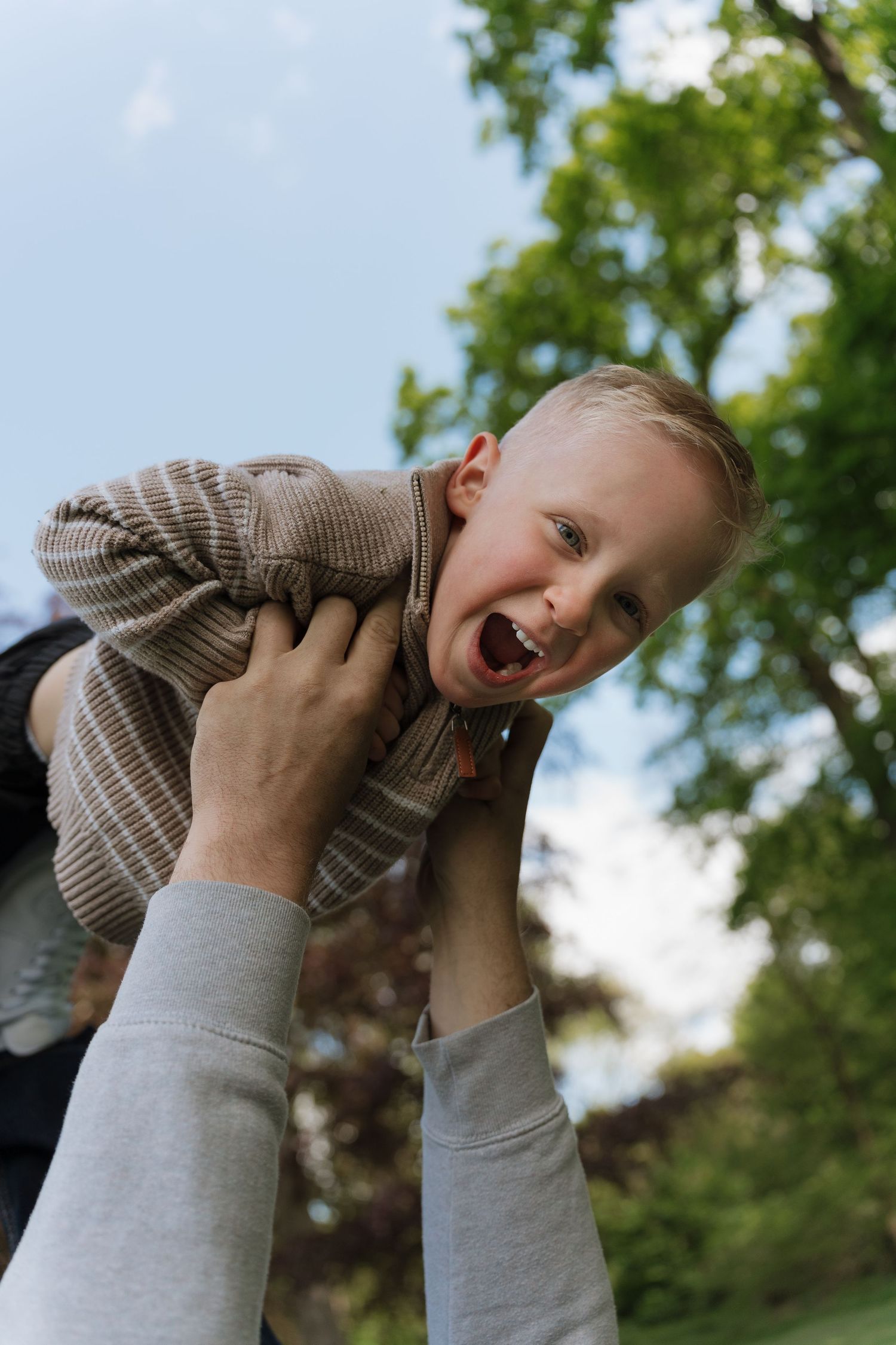 A joyful toddler boy laughing while being lifted up by an adult outdoors in a green park setting.