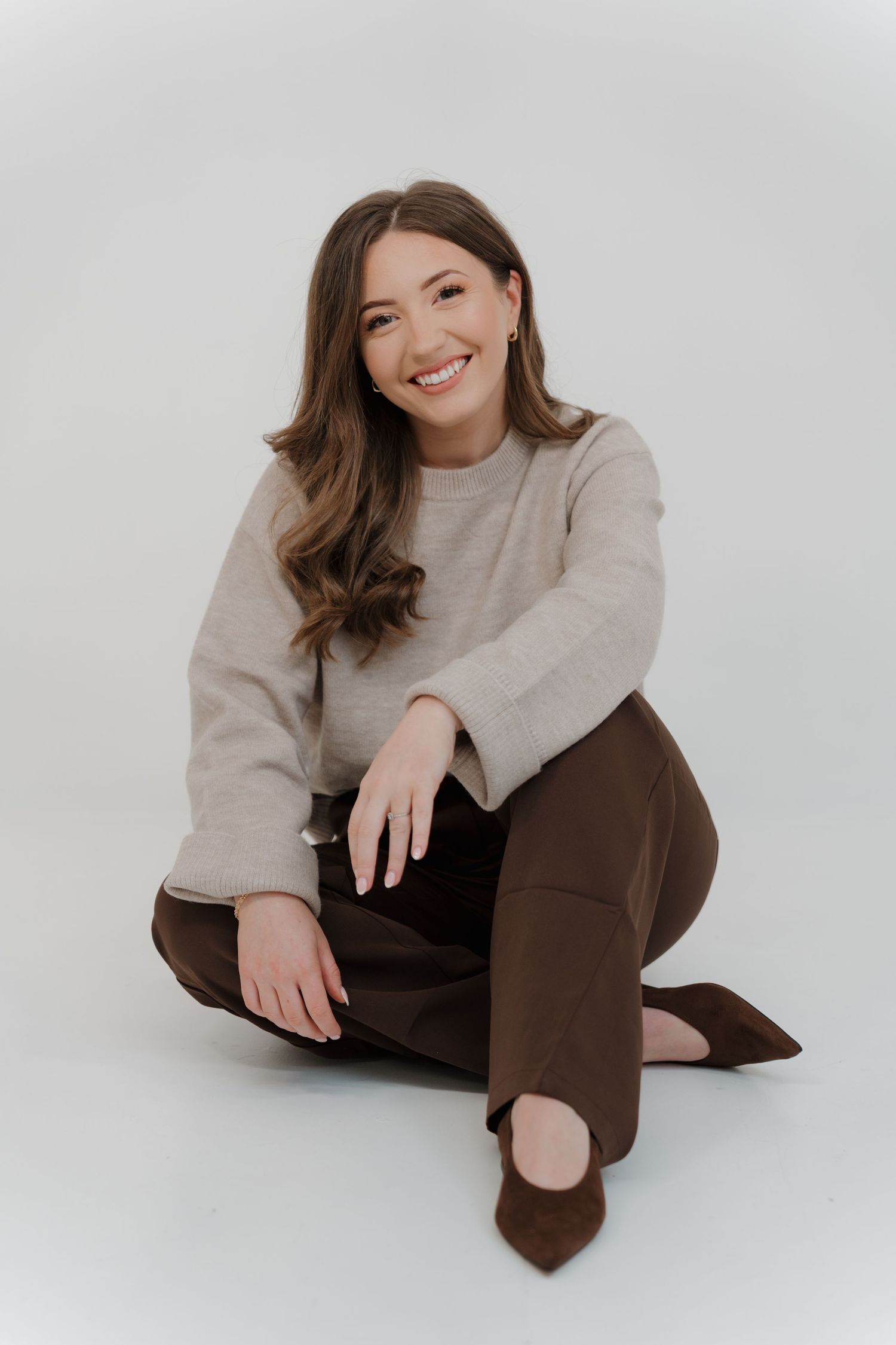 Smiling young woman in beige sweater and brown pants sitting on floor in a professional studio photo captured by Shots by Maz Photography.