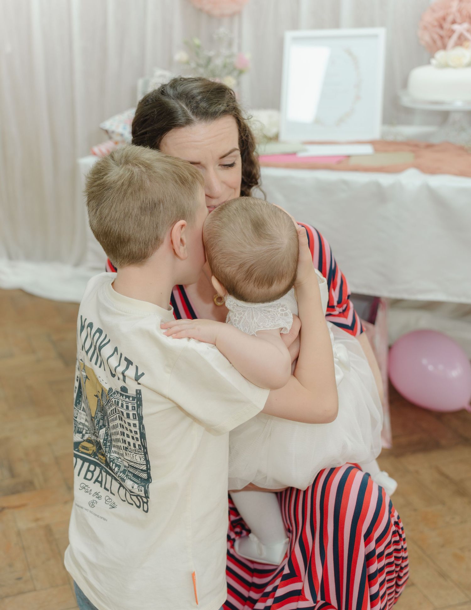 Two children share a sweet embrace while sitting on a floor decorated with pink balloons in a light-filled room.