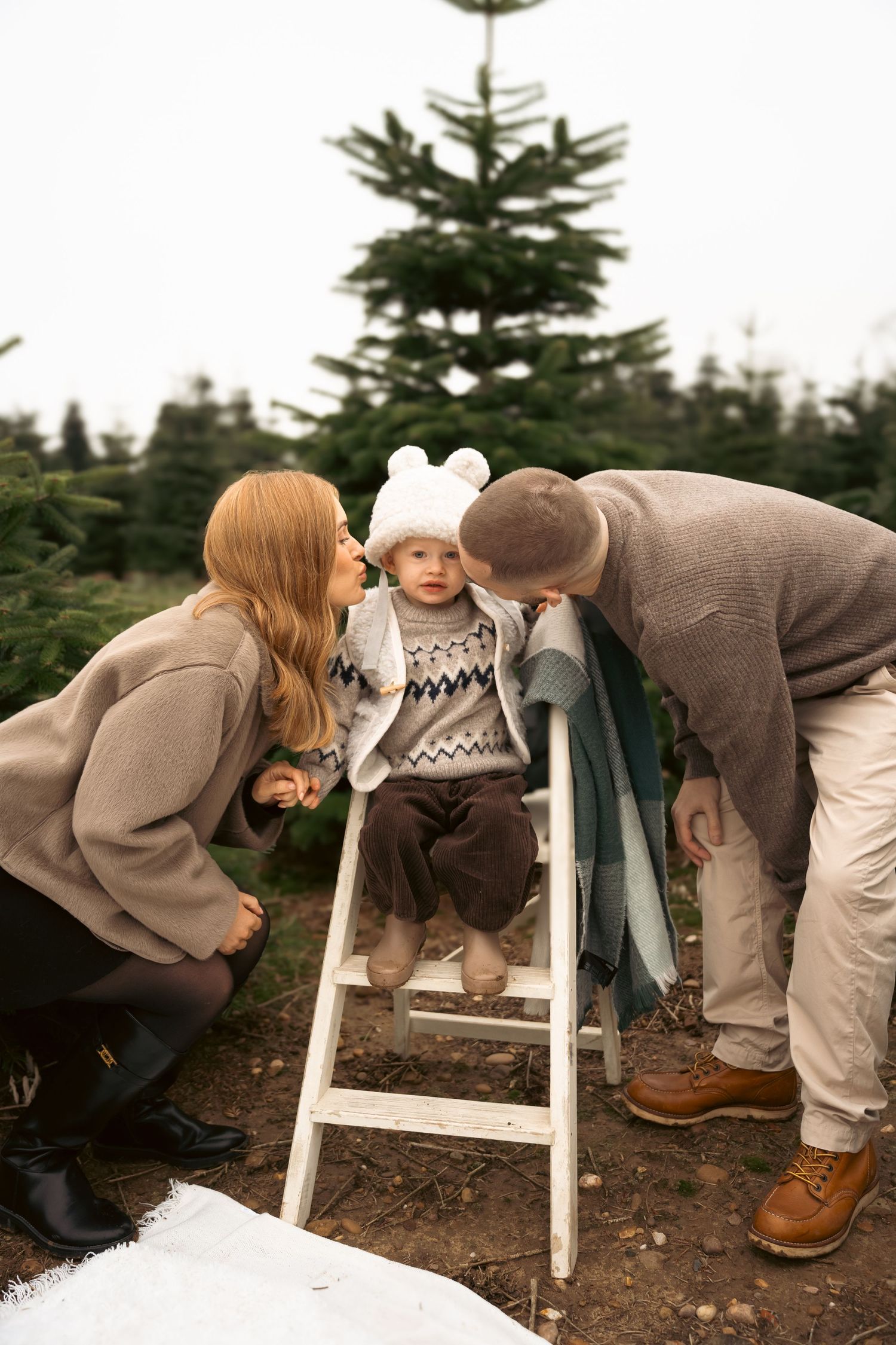 A small child in a winter sweater and hat stands on a white ladder at a Christmas tree farm while two adults lean in close.