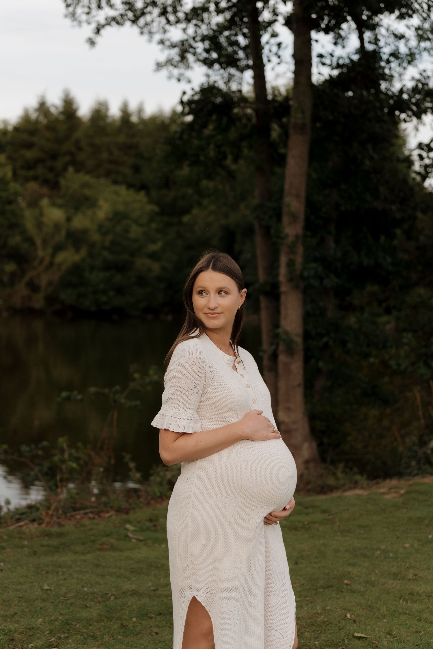 A pregnant woman in a white dress stands in a natural outdoor setting with trees and grass at sunset.
