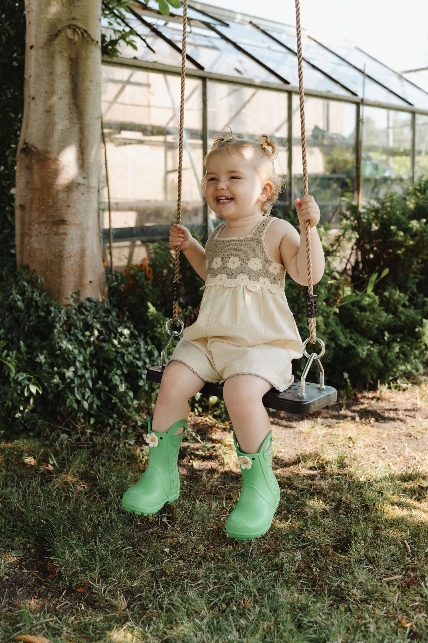 A child wearing a cream dress and bright green boots sits joyfully on a wooden swing near a greenhouse.