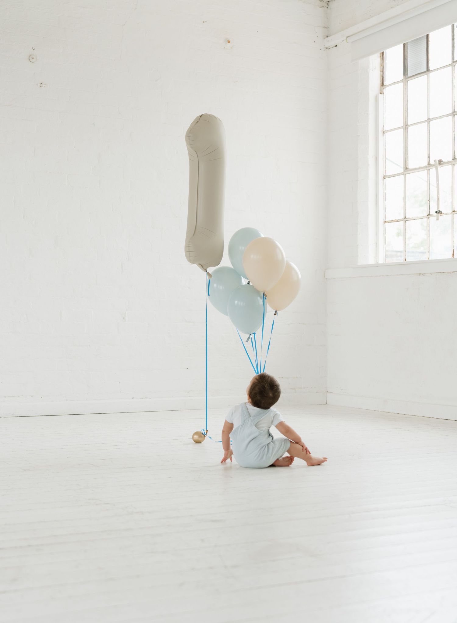 Baby sitting on white floor with number one balloon and pastel blue and cream balloons for first birthday.