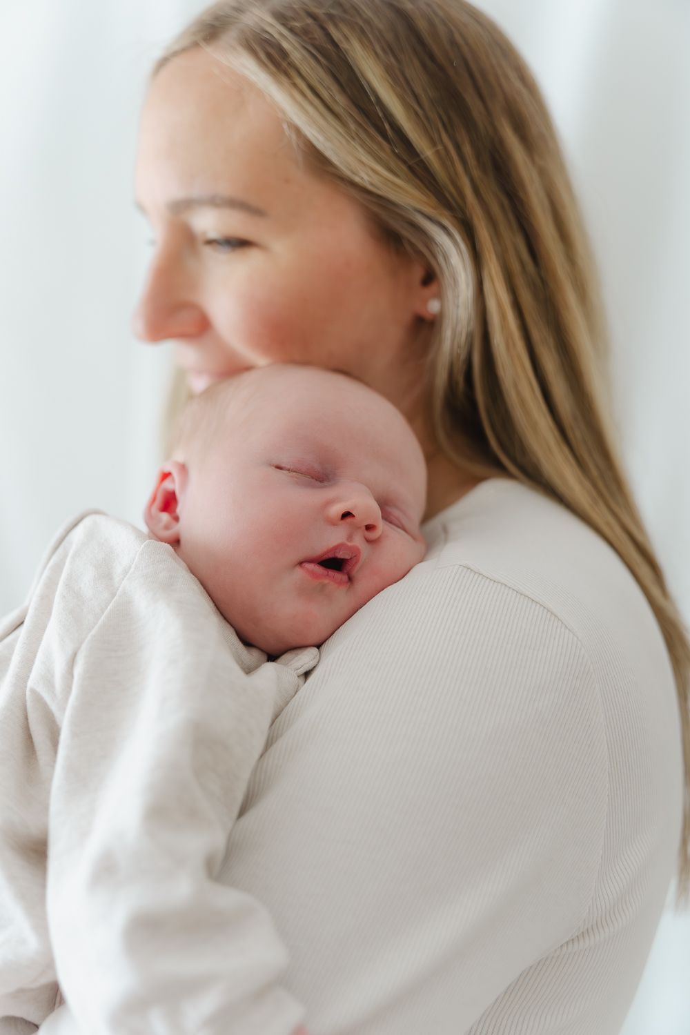A sleeping baby in white clothing rests peacefully against someone's shoulder in a bright, serene setting.