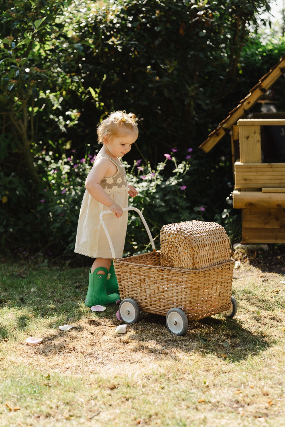 A child pushes a wicker basket cart through a sunny garden path while playing outdoors.