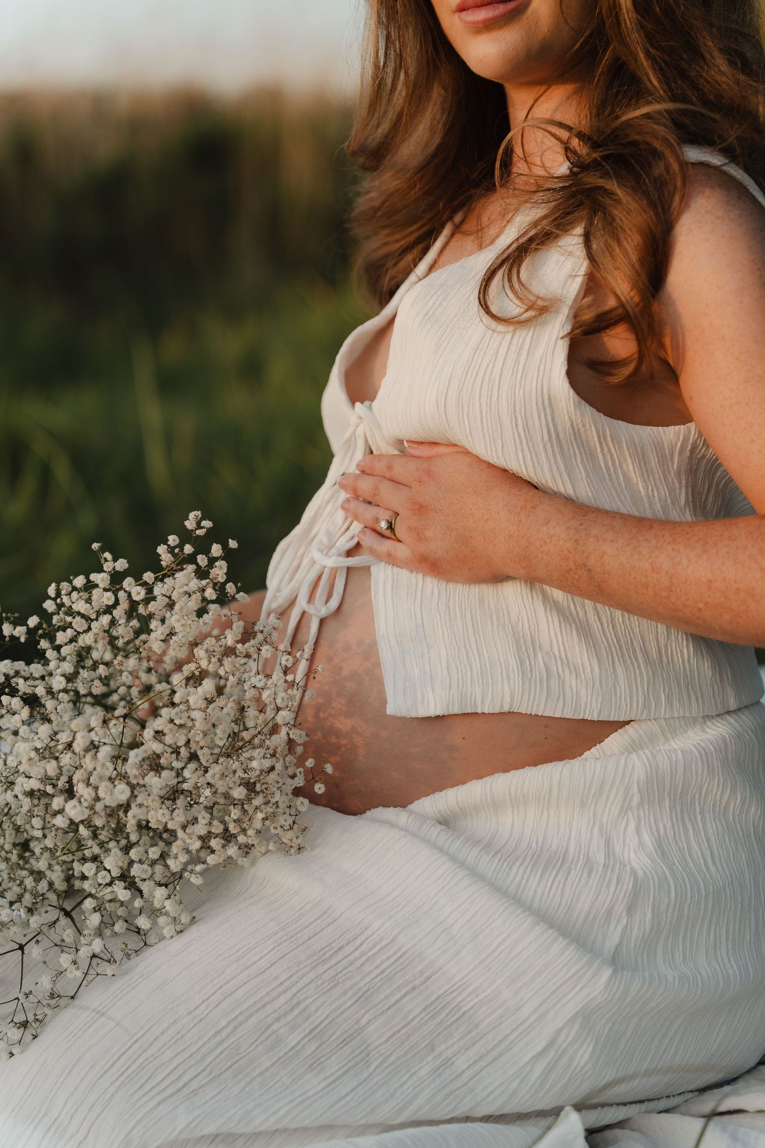 Maternity photo featuring white dress and baby's breath flowers against natural backdrop.