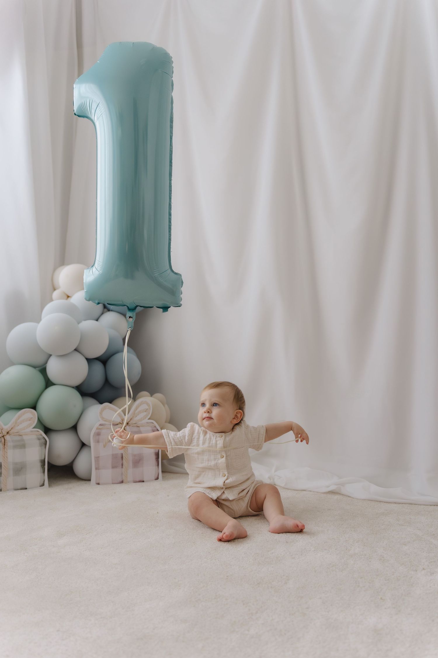 Baby crawling near large blue number one balloon and white balloons against white backdrop.