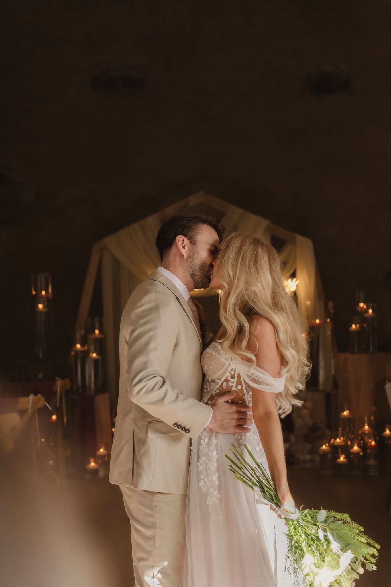 A romantic wedding couple shares an intimate dance in a dimly lit venue with glowing candles in the background.