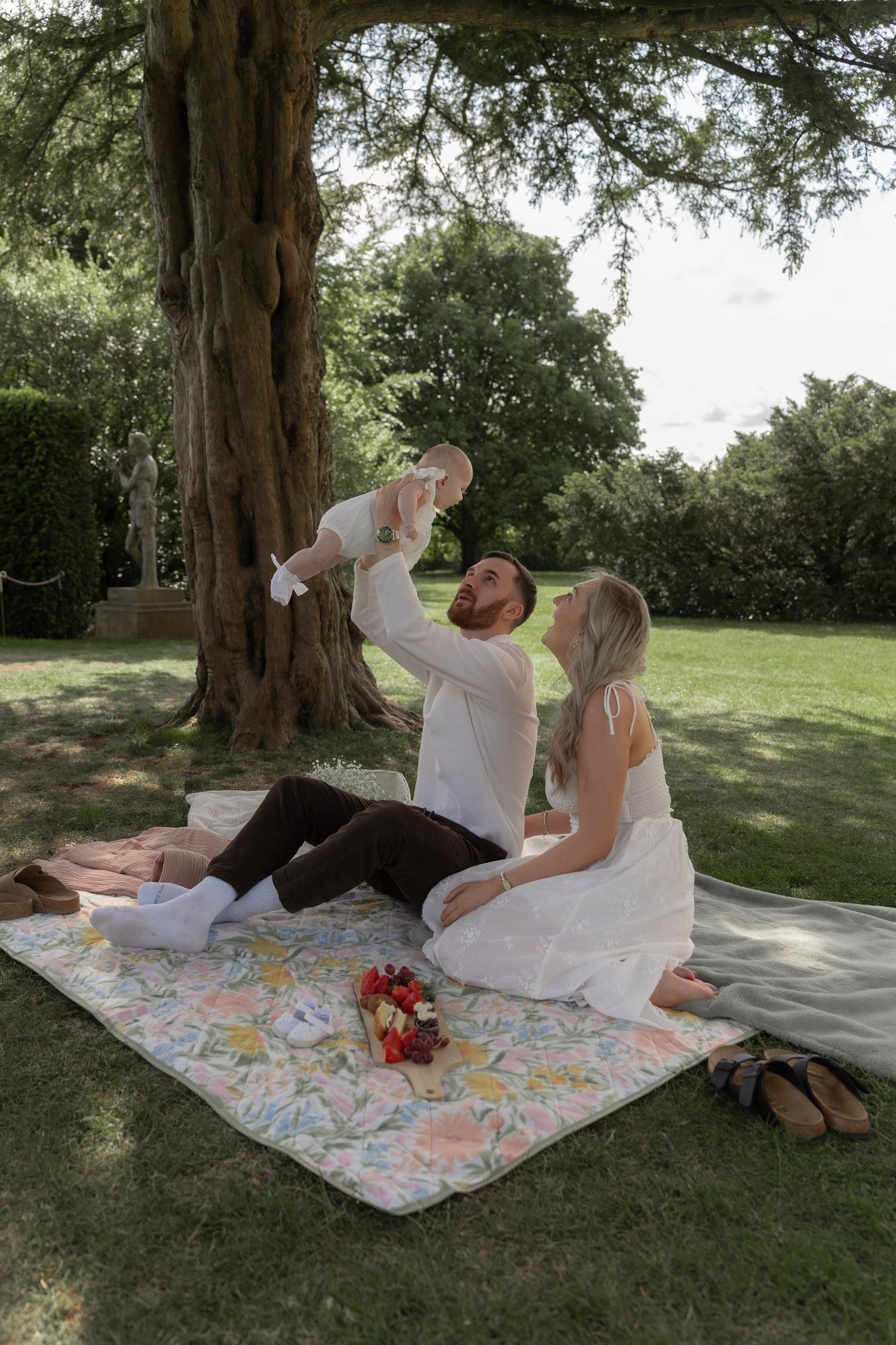 A couple enjoys a romantic summer picnic under a large tree on a floral patterned blanket.