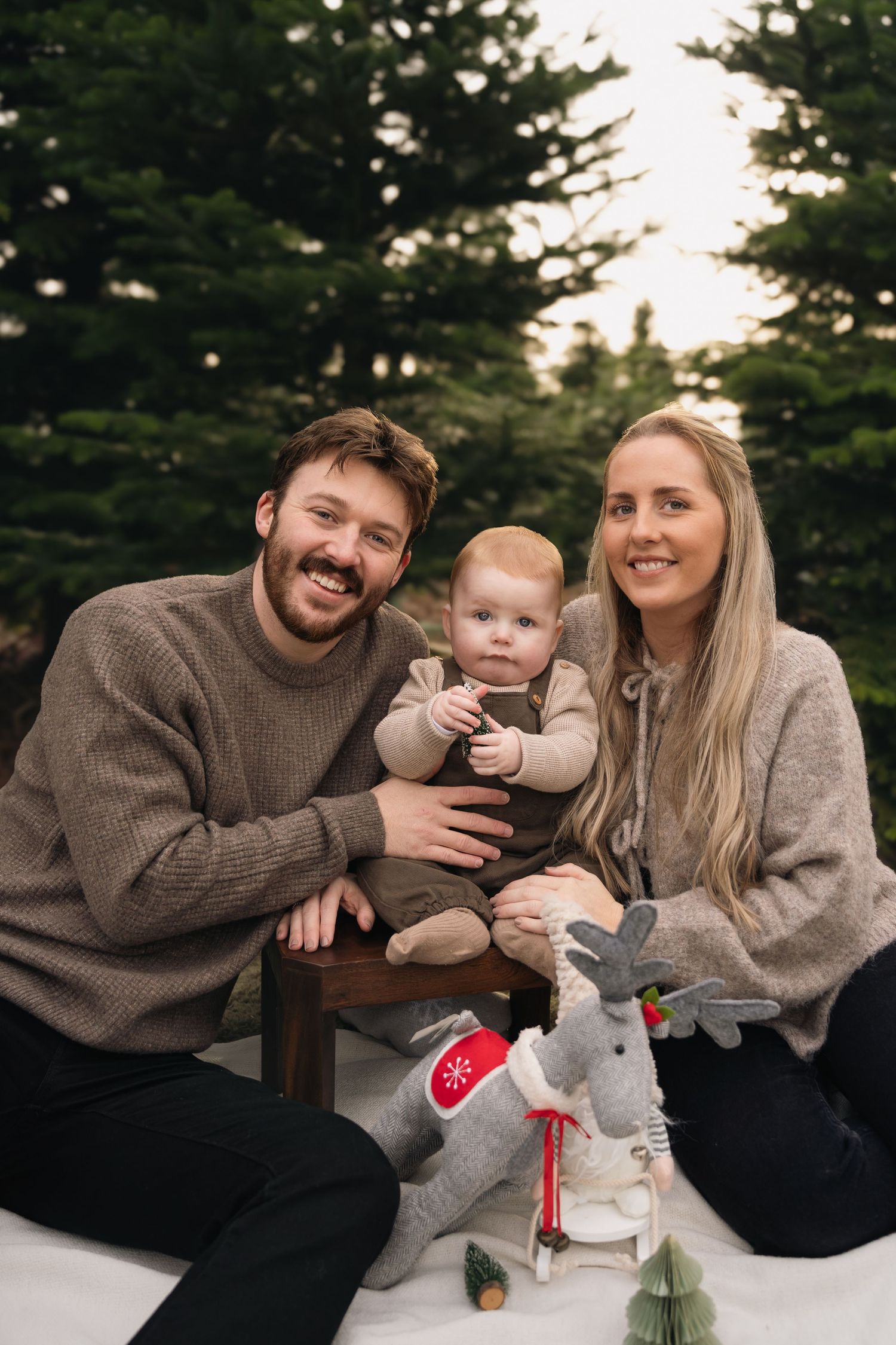 A family sits together in the snow during a winter photo session with their baby and plush reindeer toy.