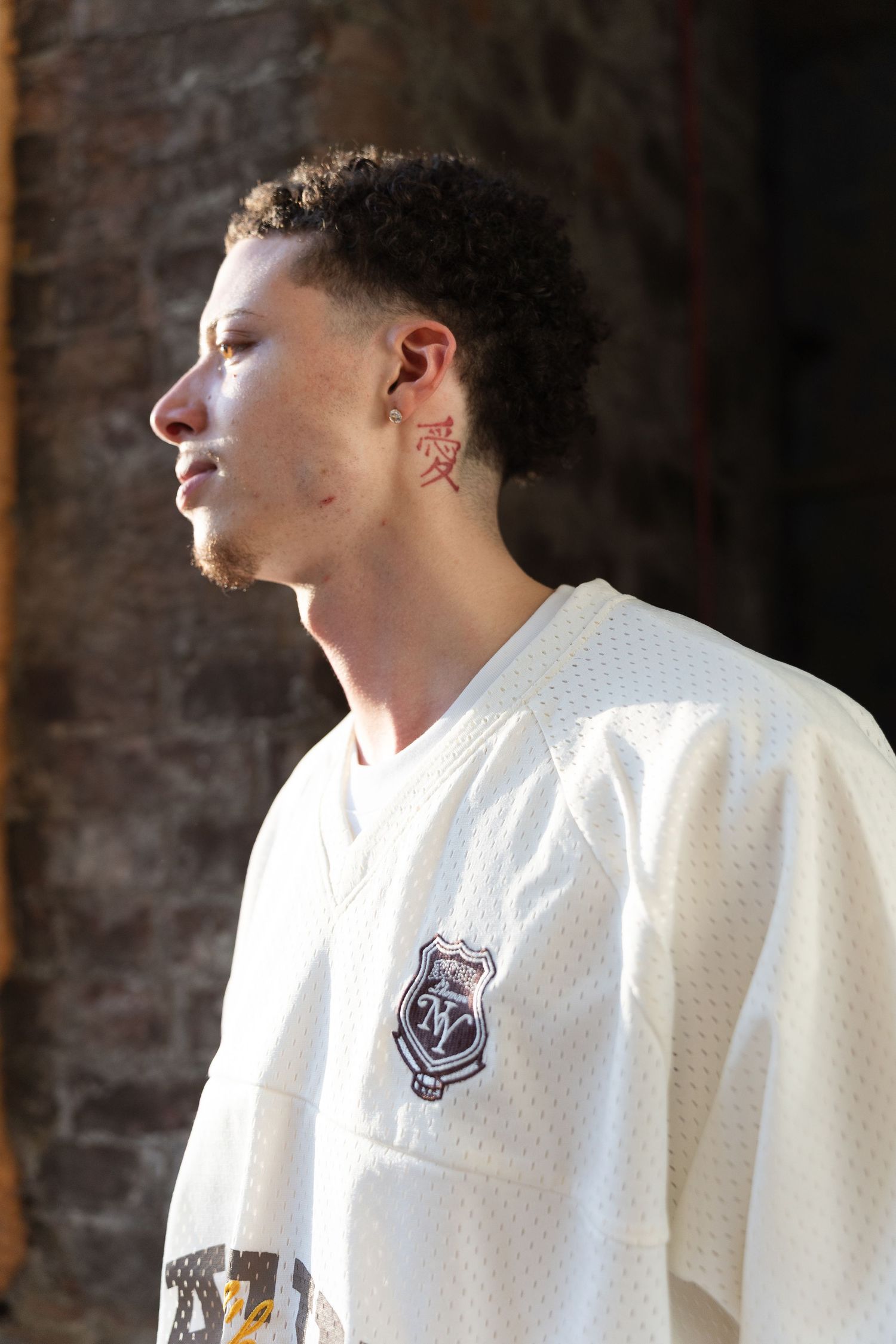 Person wearing a white jacket with embroidered patch detail shown in profile against a brick wall background.