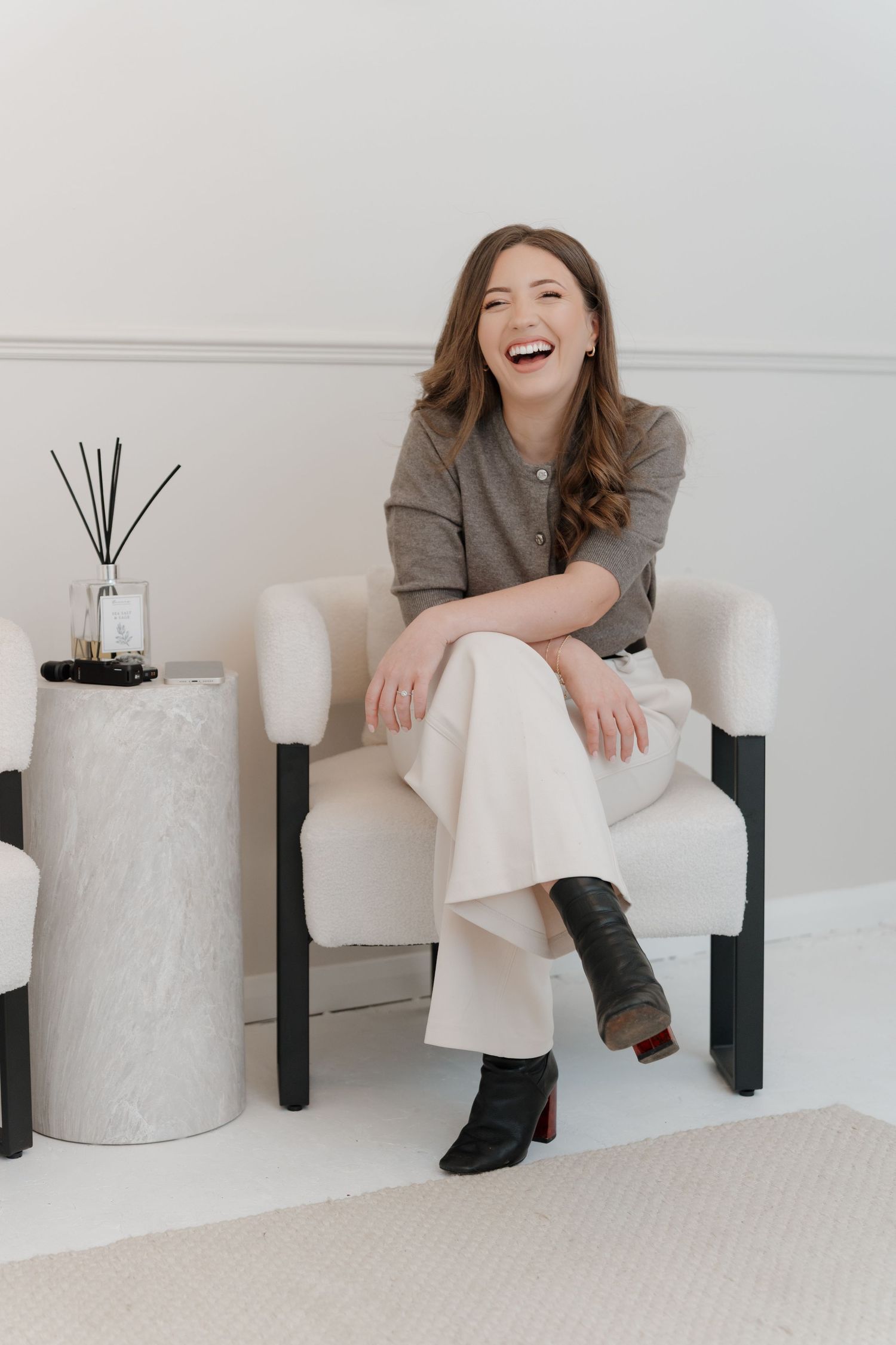 A laughing woman in white trousers and gray top sits in a white armchair beside a side table with reed diffuser.
