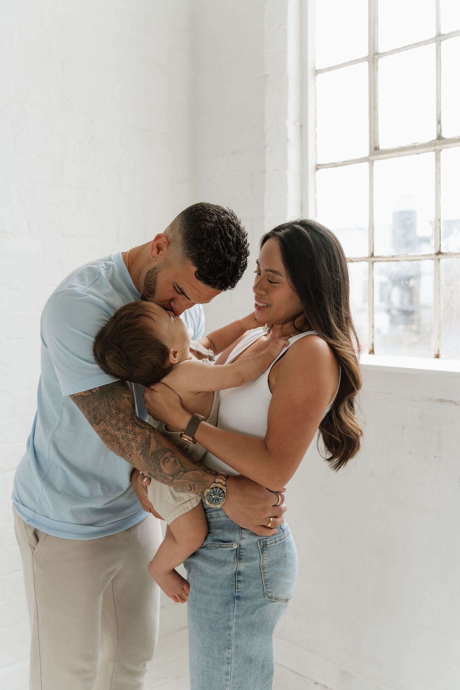 Young couple sharing a tender moment with their baby in a bright white room near a large window.