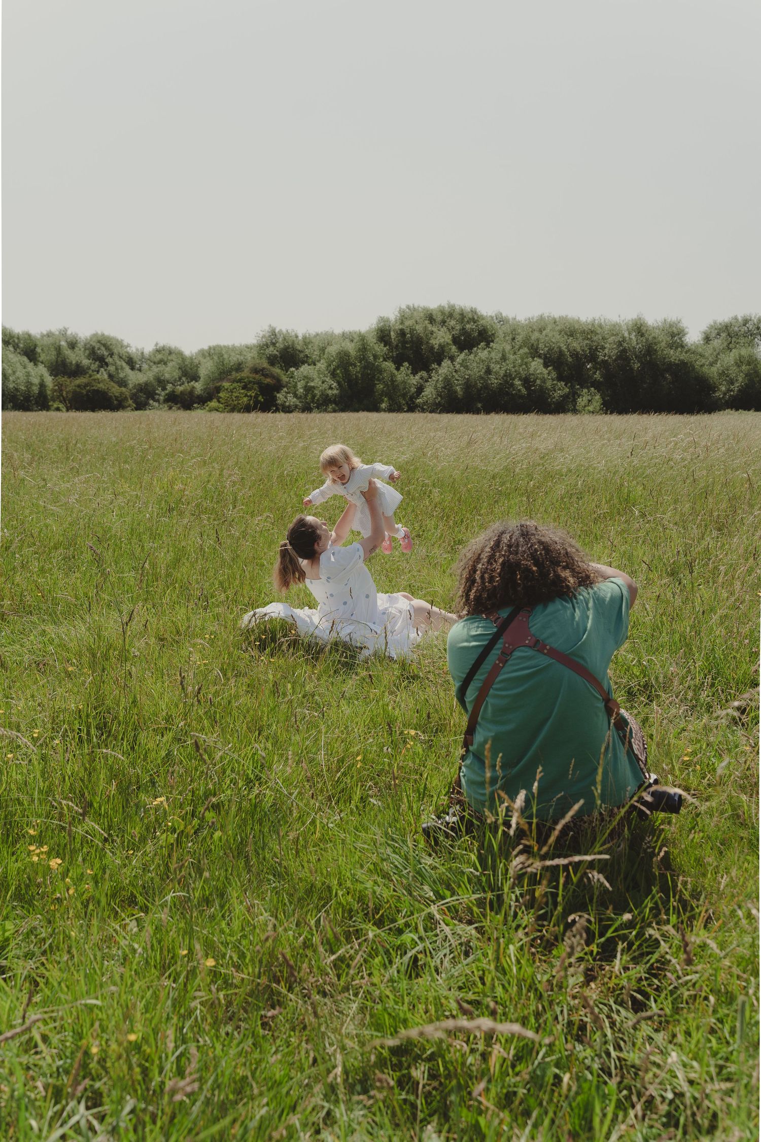 A photographer in a green shirt captures a photoshoot in a sunny meadow with tall grass and trees in the background.
